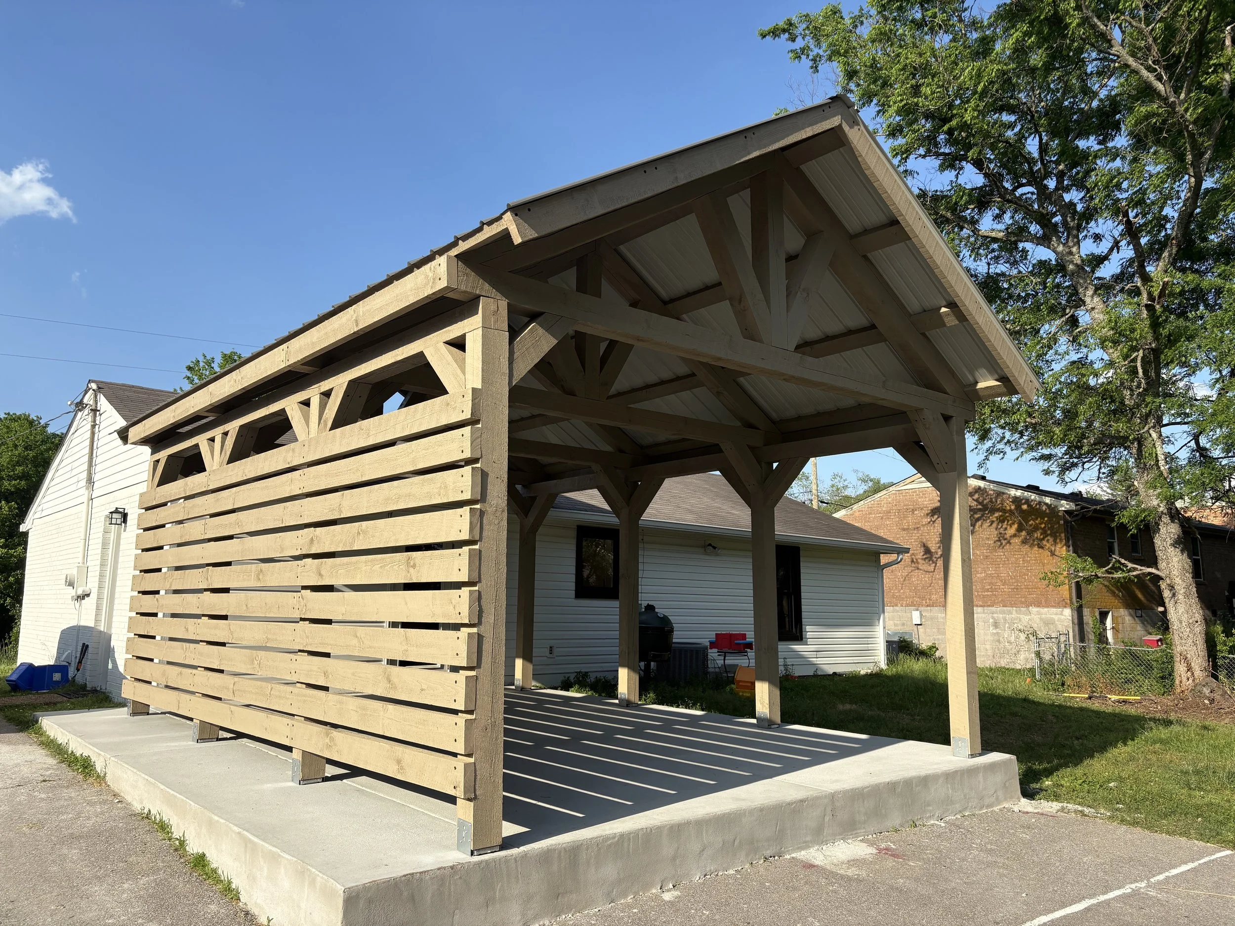 Newly constructed wooden carport with slatted side walls and a metal roof, adjacent to a white house with vinyl siding and a concrete pad foundation, under a clear blue sky with trees in the background.