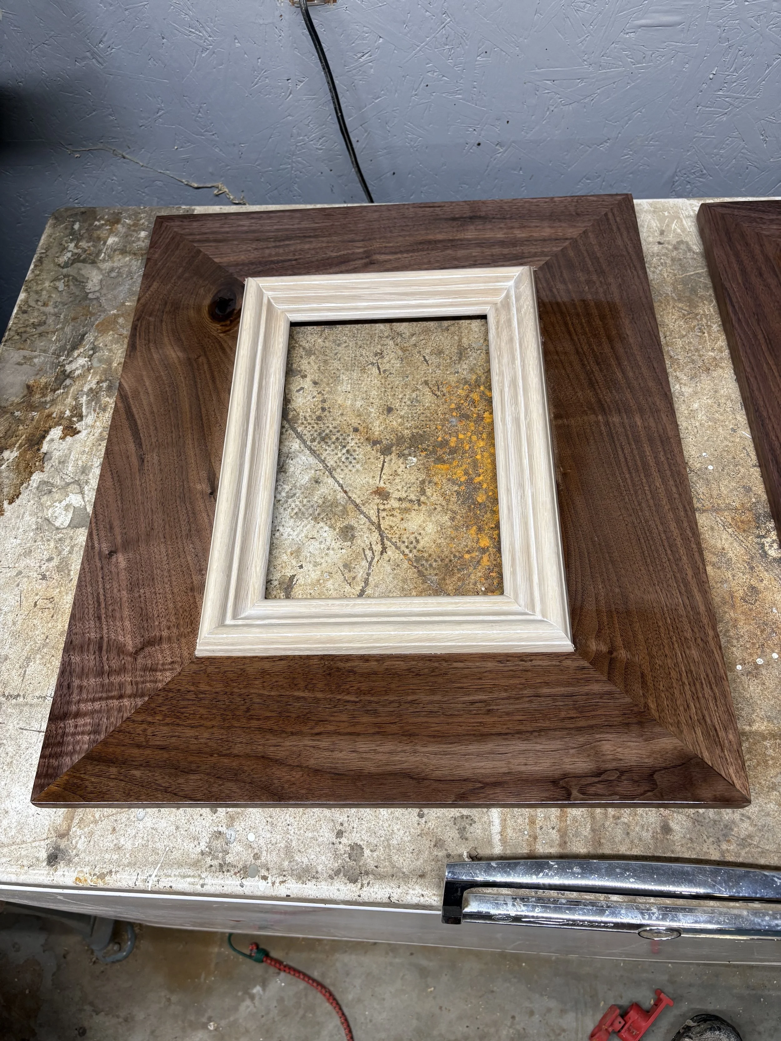 A woodworking project with a frame inside a looking glass shaped piece with a rectangular frame made of light-colored wood, placed on a workbench with a larger dark wood frame around it.
