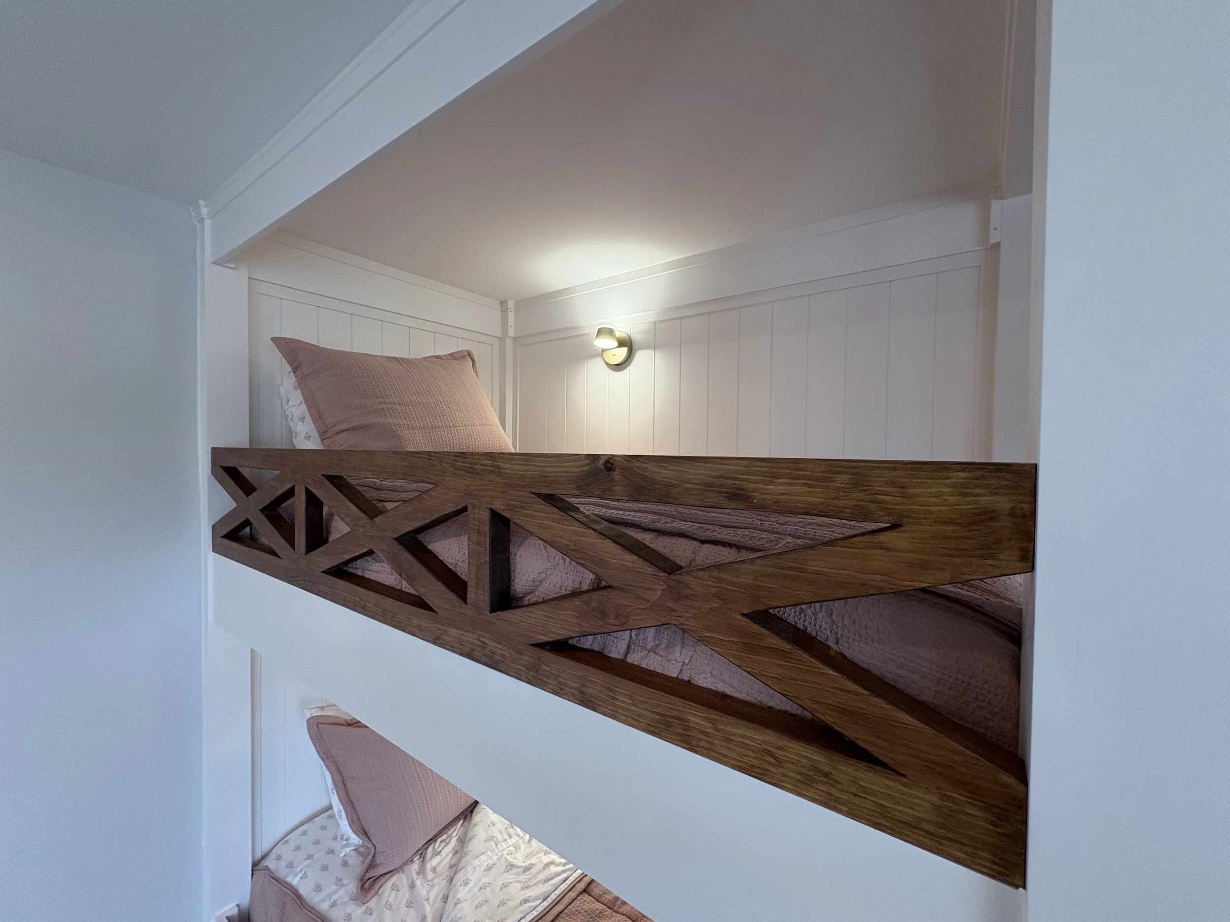 A room with white paneled walls and a wooden loft bed with a geometric design, pink bedding, and a matching pink pillow. There is a small wall sconce light fixture on the wall above the bed.