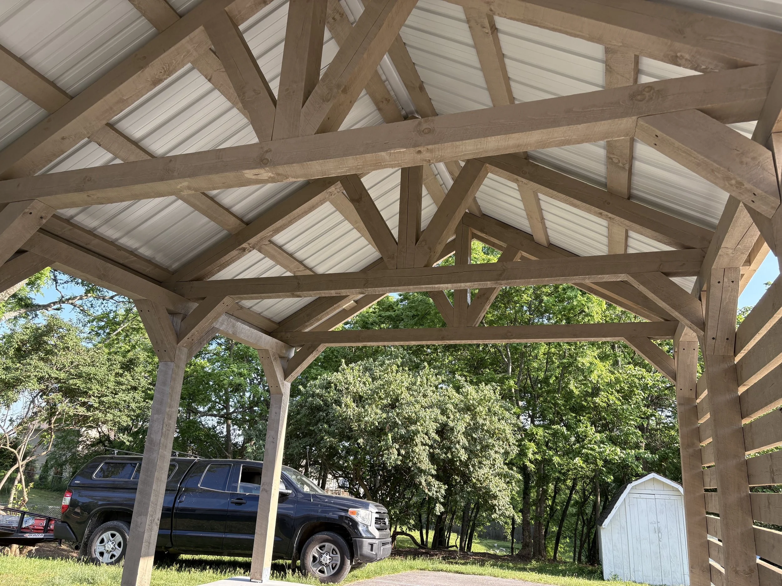 View under a wooden carport with a black truck parked underneath, surrounded by green trees and a white shed in the background.