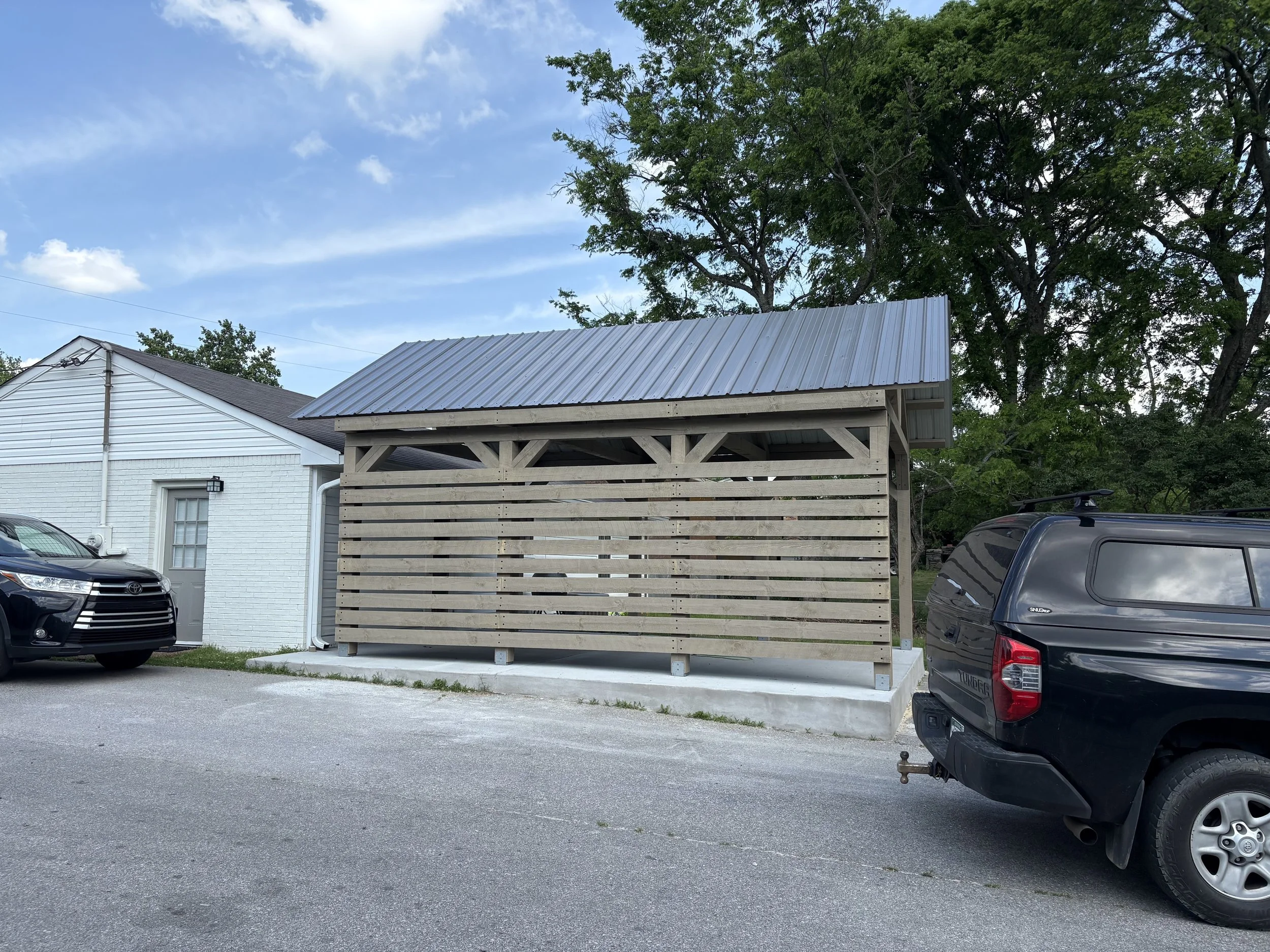 A small wooden shelter with a metal roof, situated next to a white brick building and vehicles, with trees and a blue sky in the background.