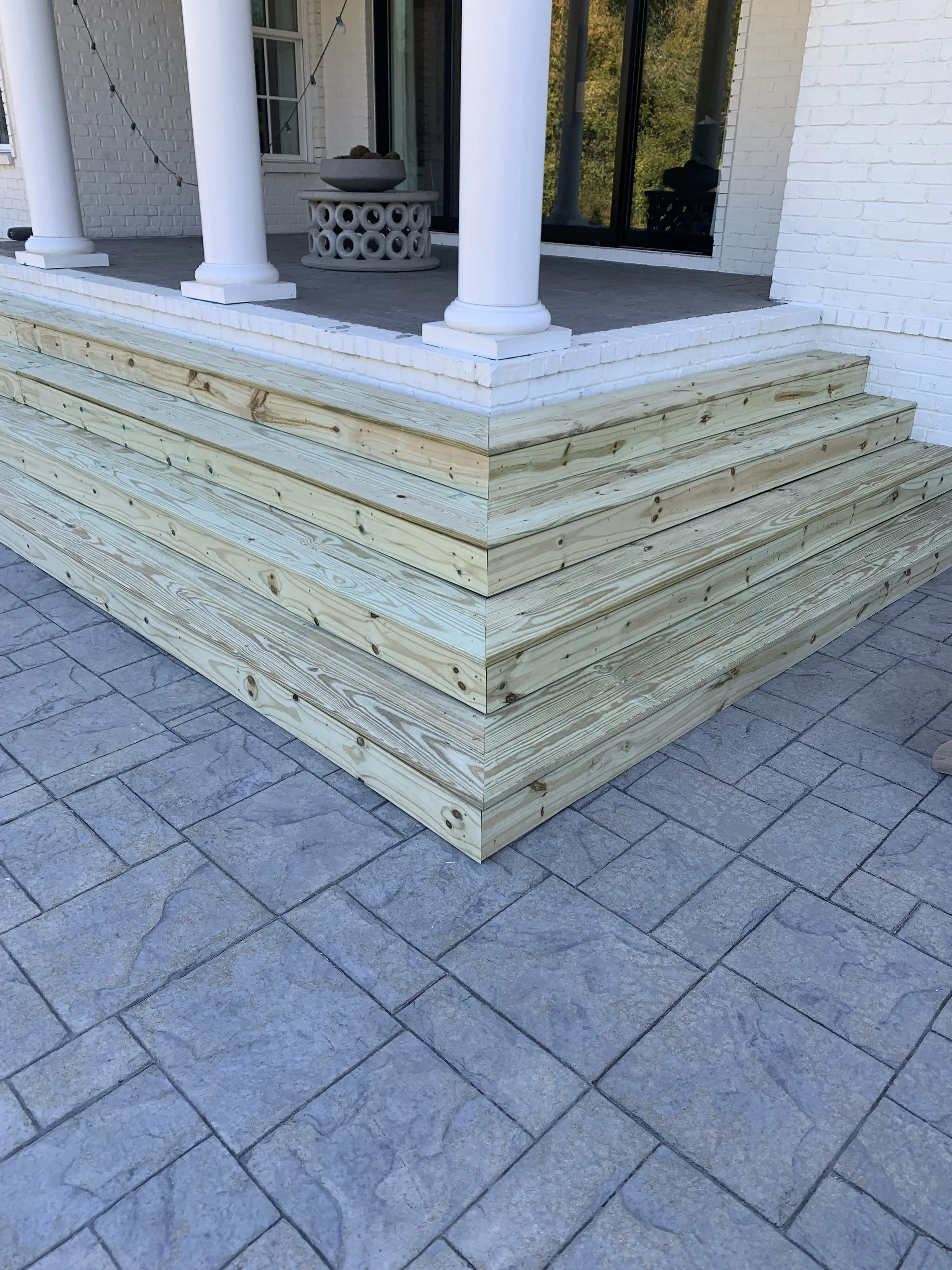 A house porch with white brick columns and railing, topped with string lights, and new wooden steps leading up to it, with a stone-paved patio below.