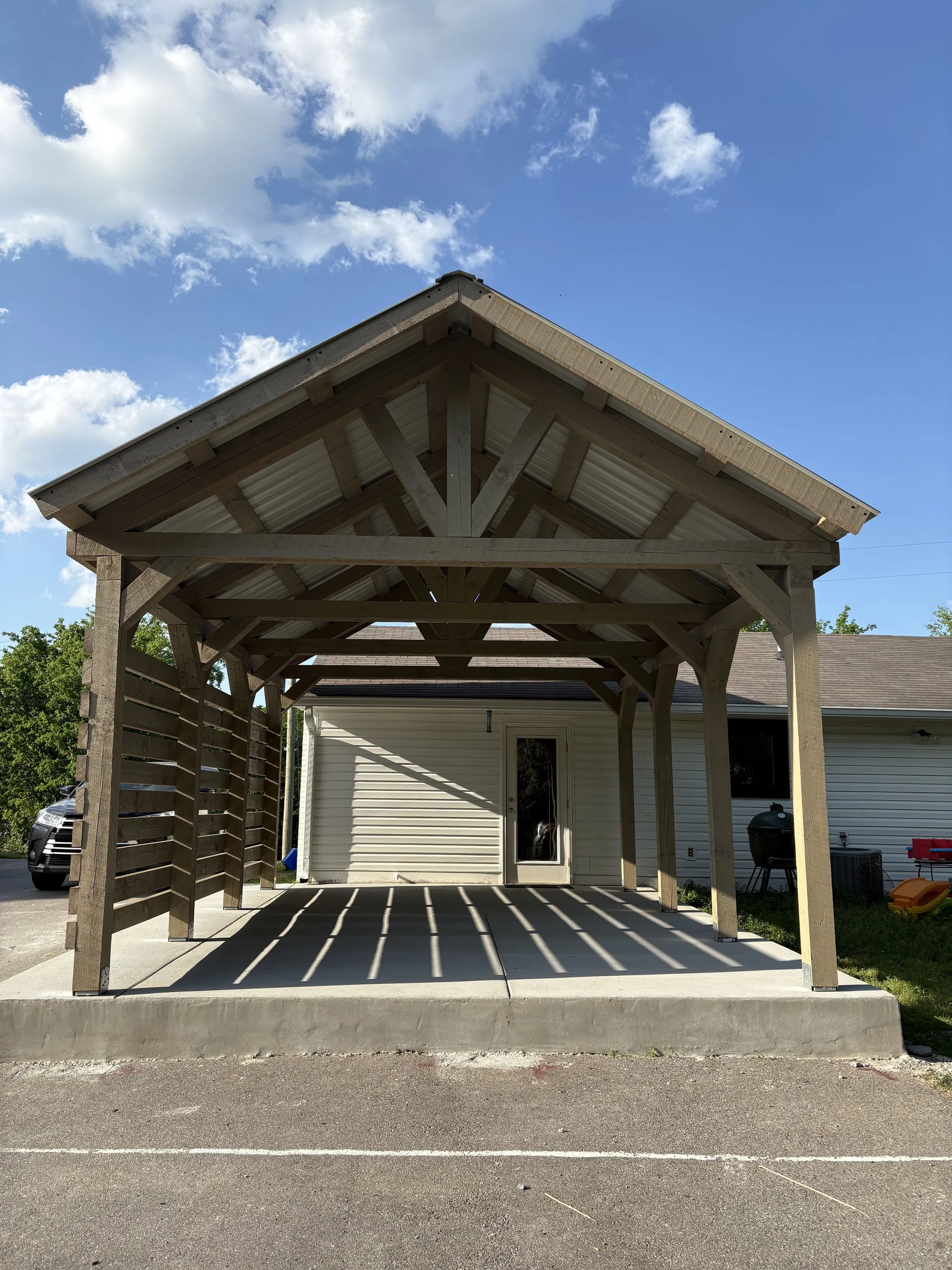 A wooden carport with a gabled roof offers parking space beneath. The structure is built with new timber and concrete flooring, attached to a white house with vinyl siding, with a door and window visible. Backyard items like a grill, trash can, and c
