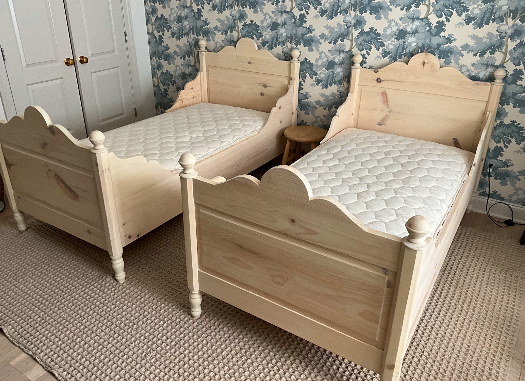 Two unfinished wooden twin beds with white mattresses, positioned parallel to each other in a bedroom with a patterned blue and white wallpaper and a beige textured rug.