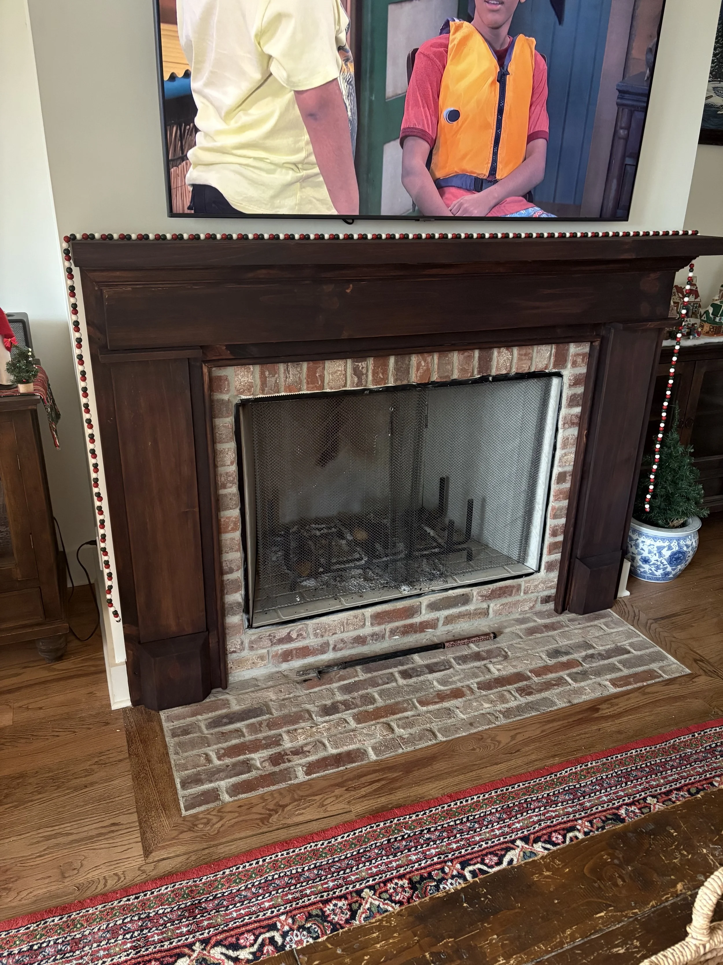 Living room fireplace with a dark wooden mantle, brick surround, and a television mounted above. The fireplace has a mesh screen, and the hearth features exposed brick. There is a decorated Christmas tree in a blue and white vase to the right of the 