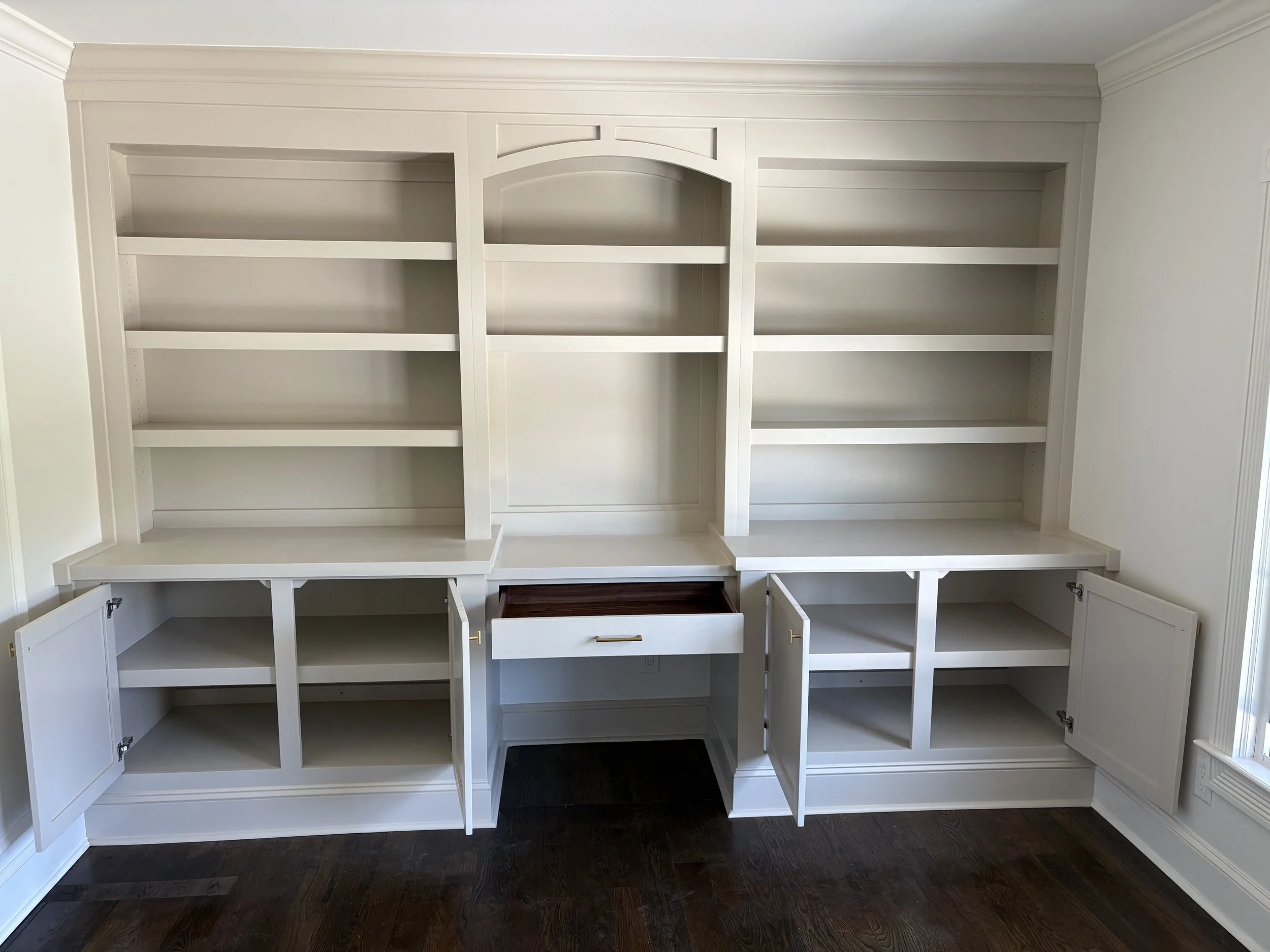 White built-in bookshelf with open cabinets and shelves, and a small drawer in the center, in a room with dark wood flooring.