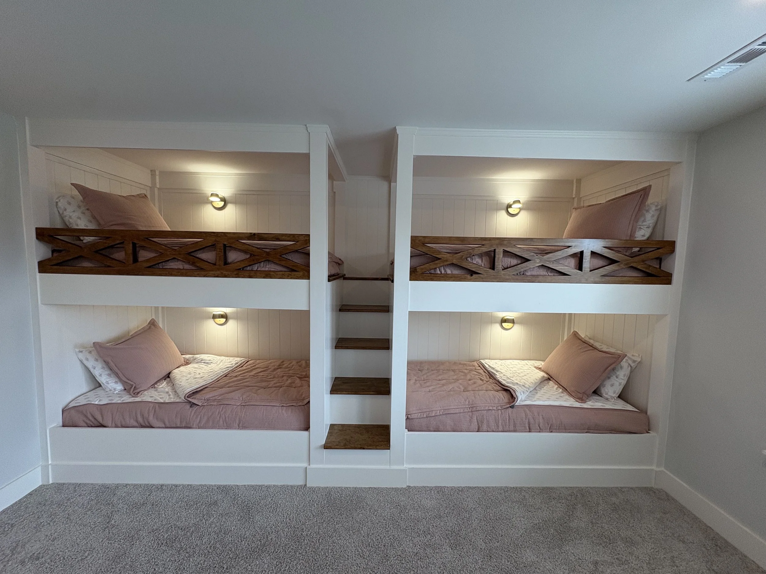 A set of four bunk beds with light brown and white linens, two on the bottom and two on the top, separated by a small wooden staircase, with wall-mounted lights above each bed, against a light-colored wall in a room with gray carpet.