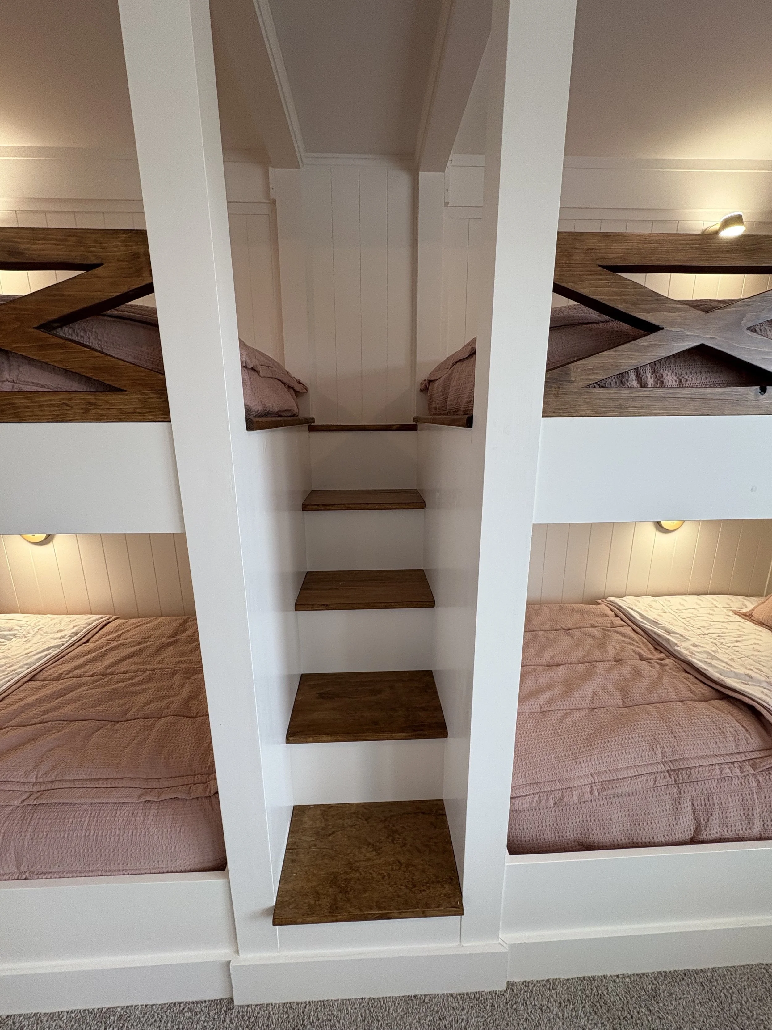 A narrow staircase with dark wood steps and white sides, situated between two sets of bunk beds with pink bedding inside a cozy room.