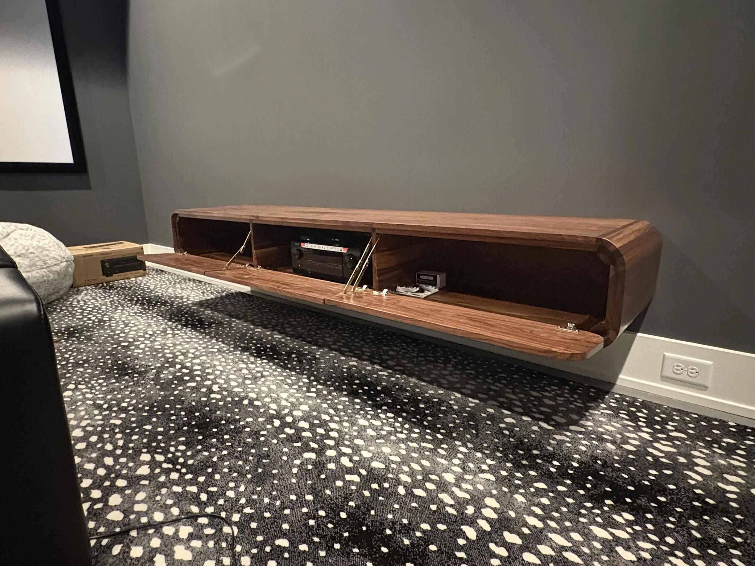 A modern wooden wall-mounted shelf with angled supports, placed above a patterned carpeted floor, with various electronic devices and objects inside the shelf.