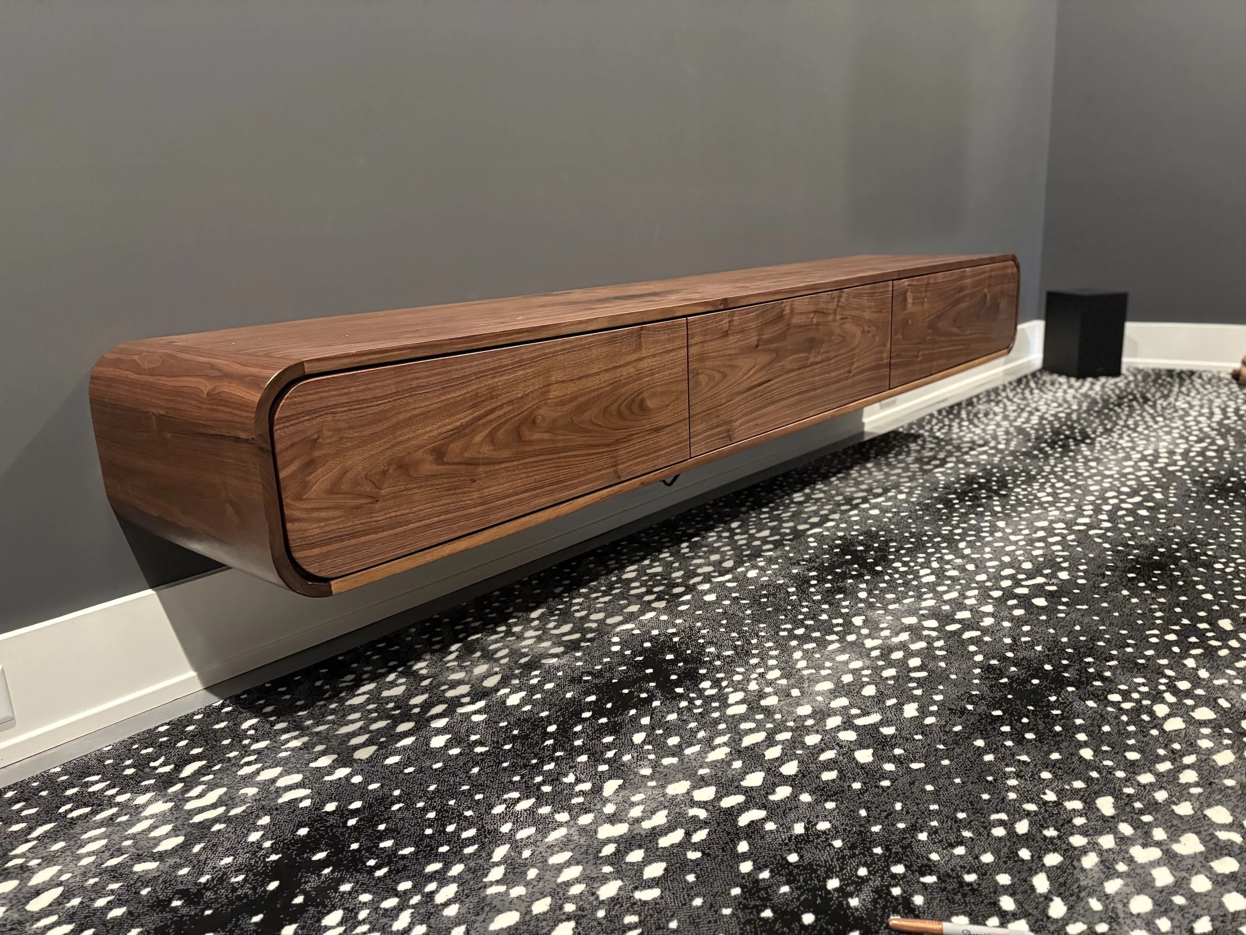 Wooden wall-mounted cabinet with rounded edges hanging above a patterned carpeted floor in a room with gray walls.