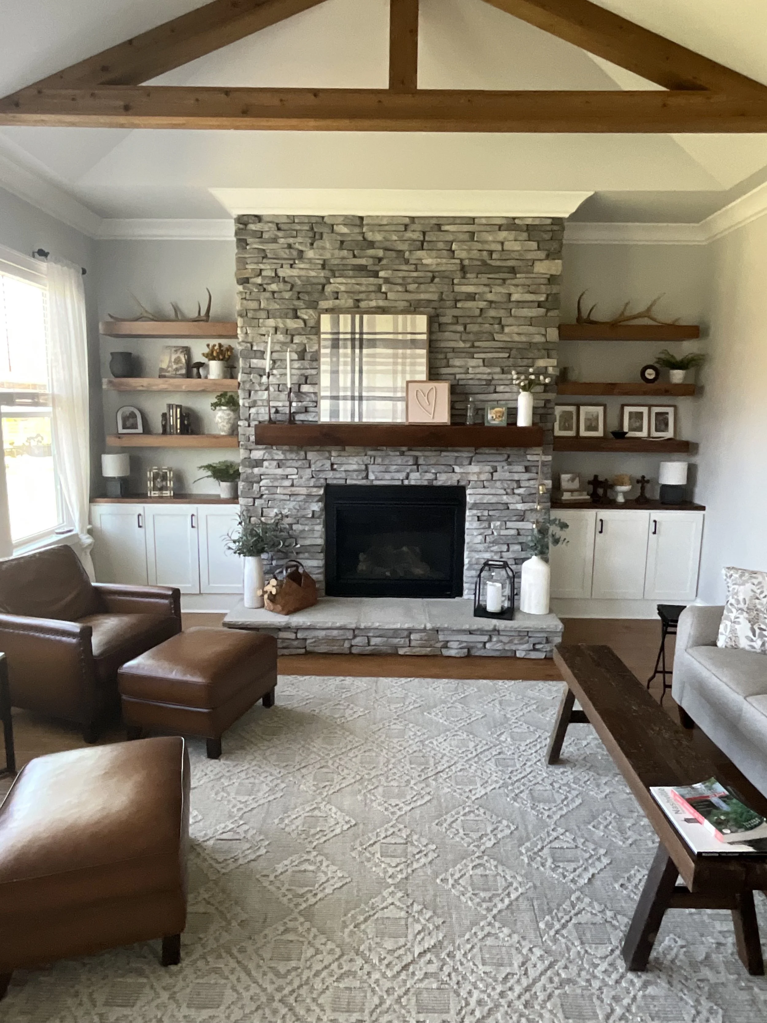 Living room with stone fireplace, wooden mantel, built-in shelves with decor, leather chairs, wooden coffee table, and a patterned rug.