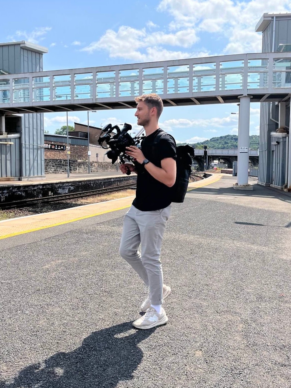 Filmmaker filming at a train station with a pedestrian bridge in the background.