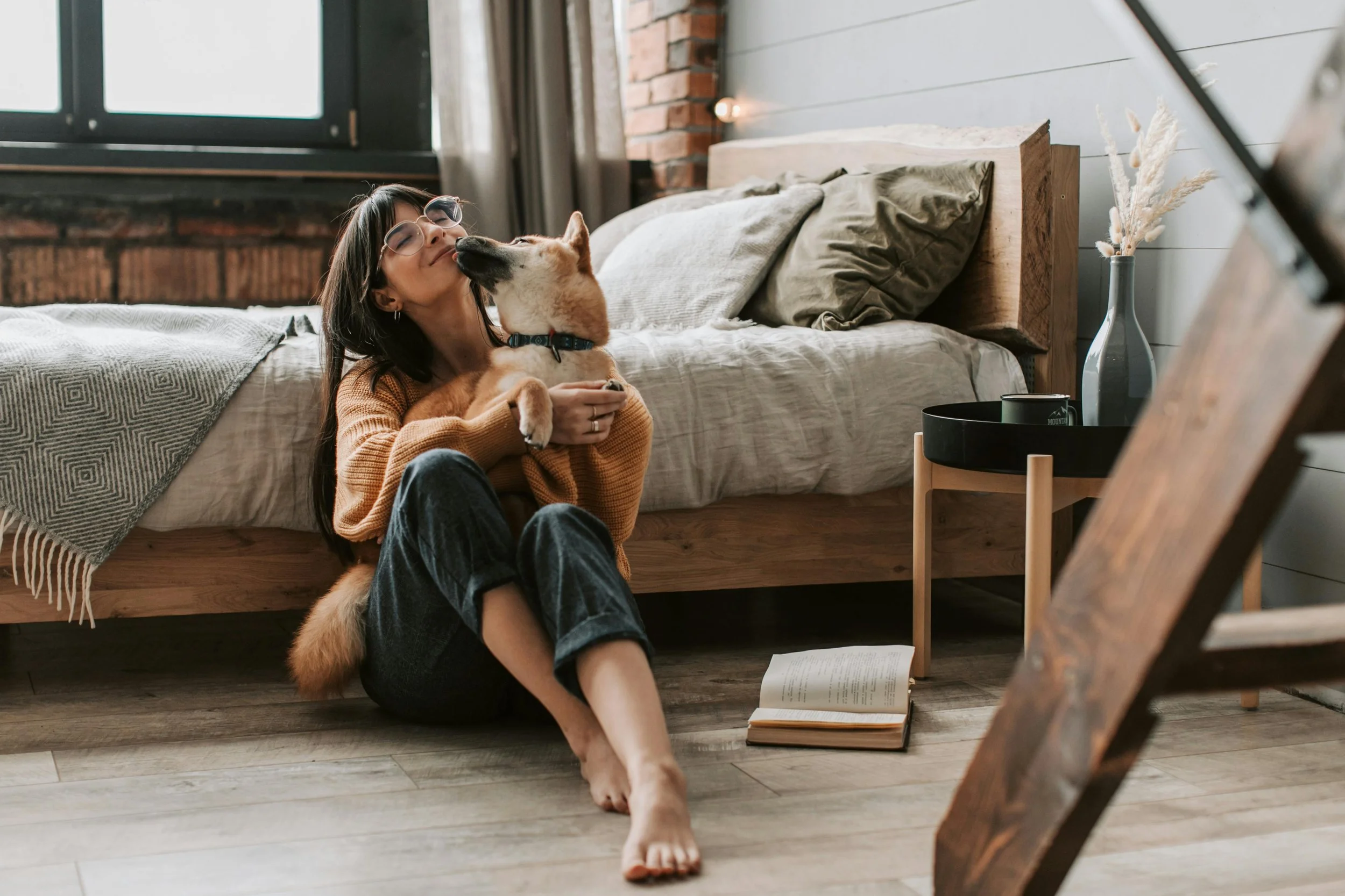 A woman is sitting on the floor in front of a bed, hugging and kissing her dog. The room has a wooden bed frame, pillows, a blanket, an open book on the floor, and a small table with a vase of dried flowers.