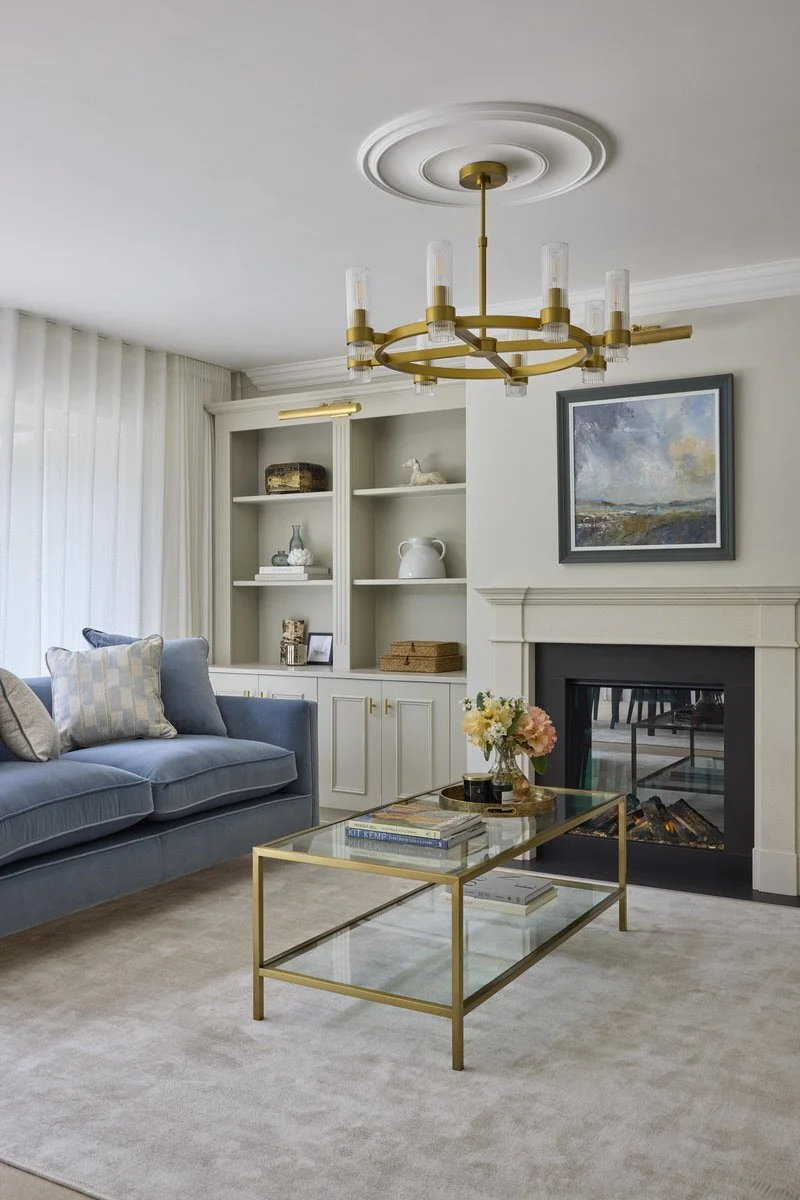 An elegant living room with a soft blue velvet sofa, brass and glass contemporary chandelier, built in soft greige shelving, glass fronted fireplace and a brass framed glass coffee table on a neutral rug.