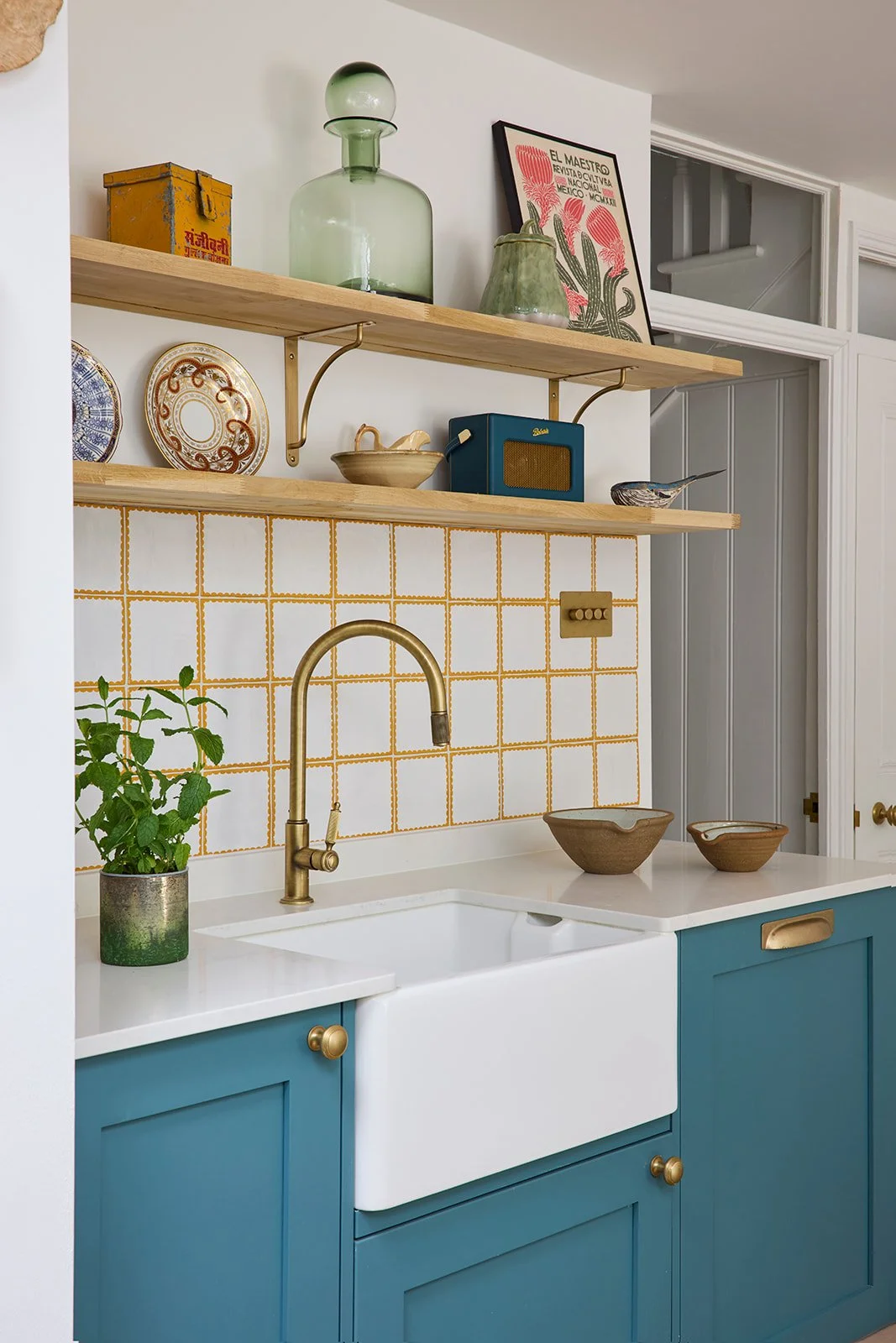 Farmhouse sink with brass finishings in a modern kitchen with teal cabinets and white and mustard yellow tile backsplash with wooden shelves