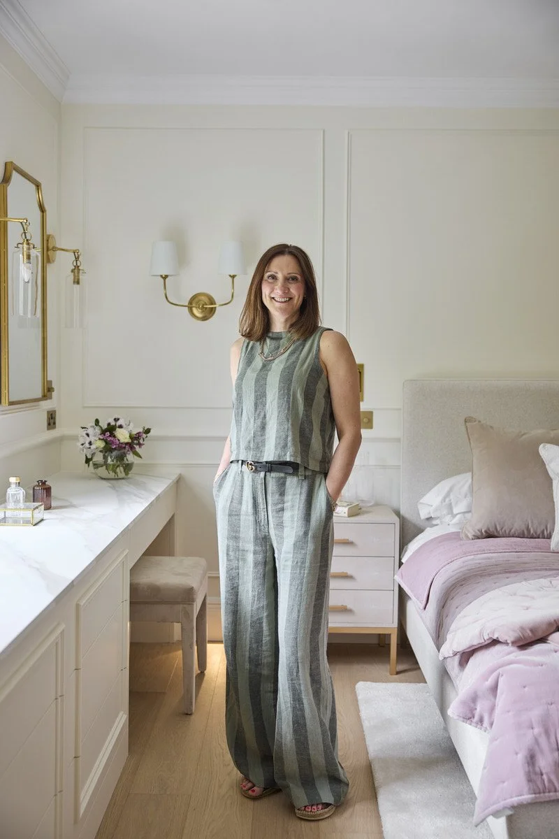 Interior designer Tracy Duncan stands smiling in an elegant cream bedroom featuring panelled walls, brass wall lights, a marble topped dressing table and a bed dressed in soft blush pink bedding.