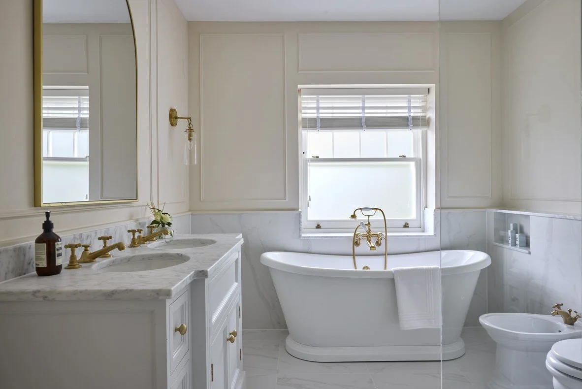 A classic bathroom with a freestanding roll-top bath, marblesurfaces, brass fixtures, panelled cream walls, and a gold-framed archmirror above a double vanity.