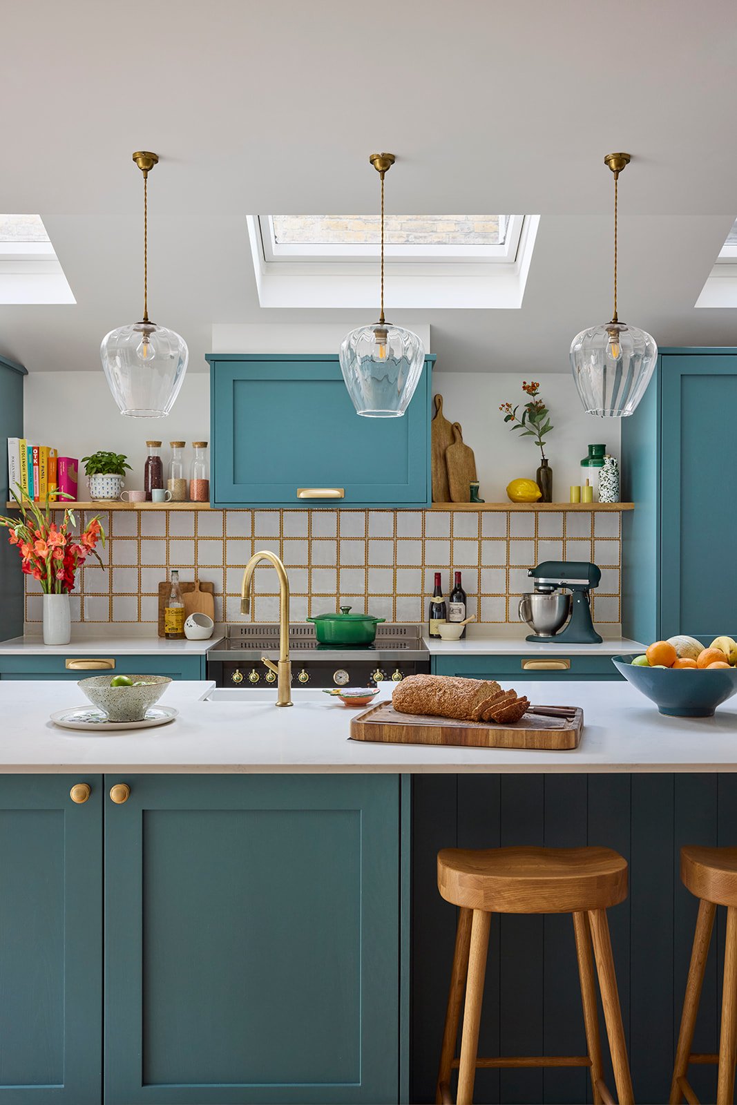 A modern kitchen with teal cabinets, a black electric stove with a green crock pot on top, white and mustard yellow tiled backsplash, and decorative items like cutting boards, jars, and plants. A farmhouse sink with brass finishings