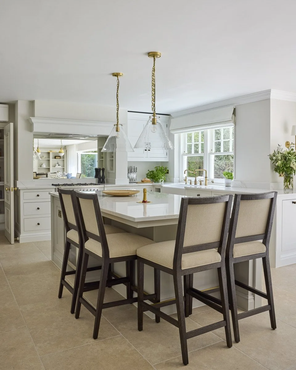 A light and airy, classic kitchen with a large marble island, four upholstered bar stools, brass chain pendant lights, and cream cabinetry on limestone tiled flooring