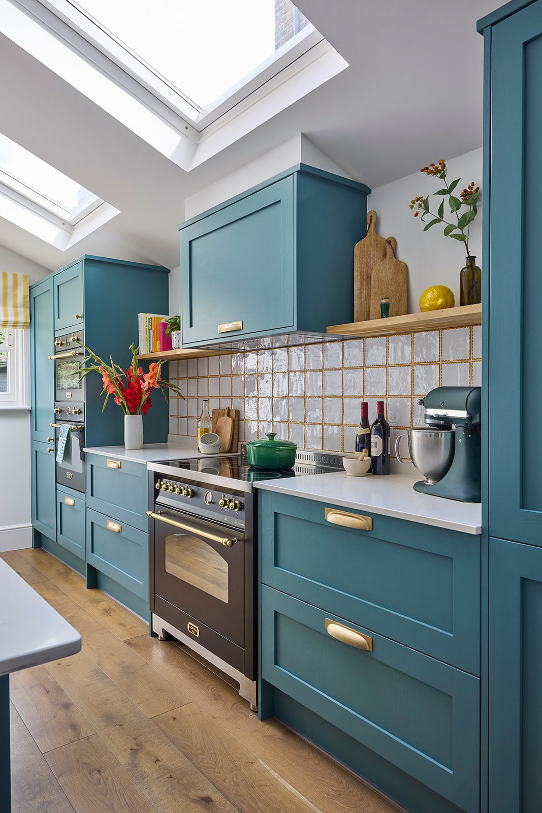 A modern kitchen with teal cabinets, a black electric stove with a green crock pot on top, white and mustard yellow tiled backsplash, and decorative items like cutting boards, jars, and plants.