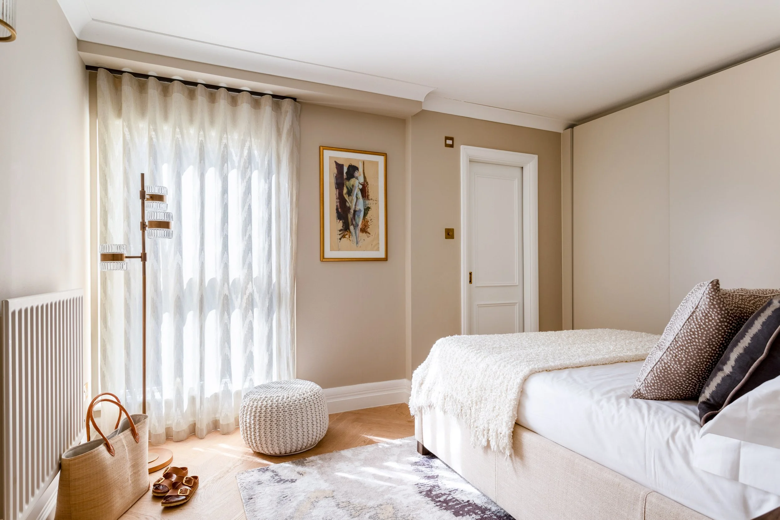 Small apartment bedroom in soft neutral tones with sheer curtains, upholstered bed and knitted pouf to maximise light