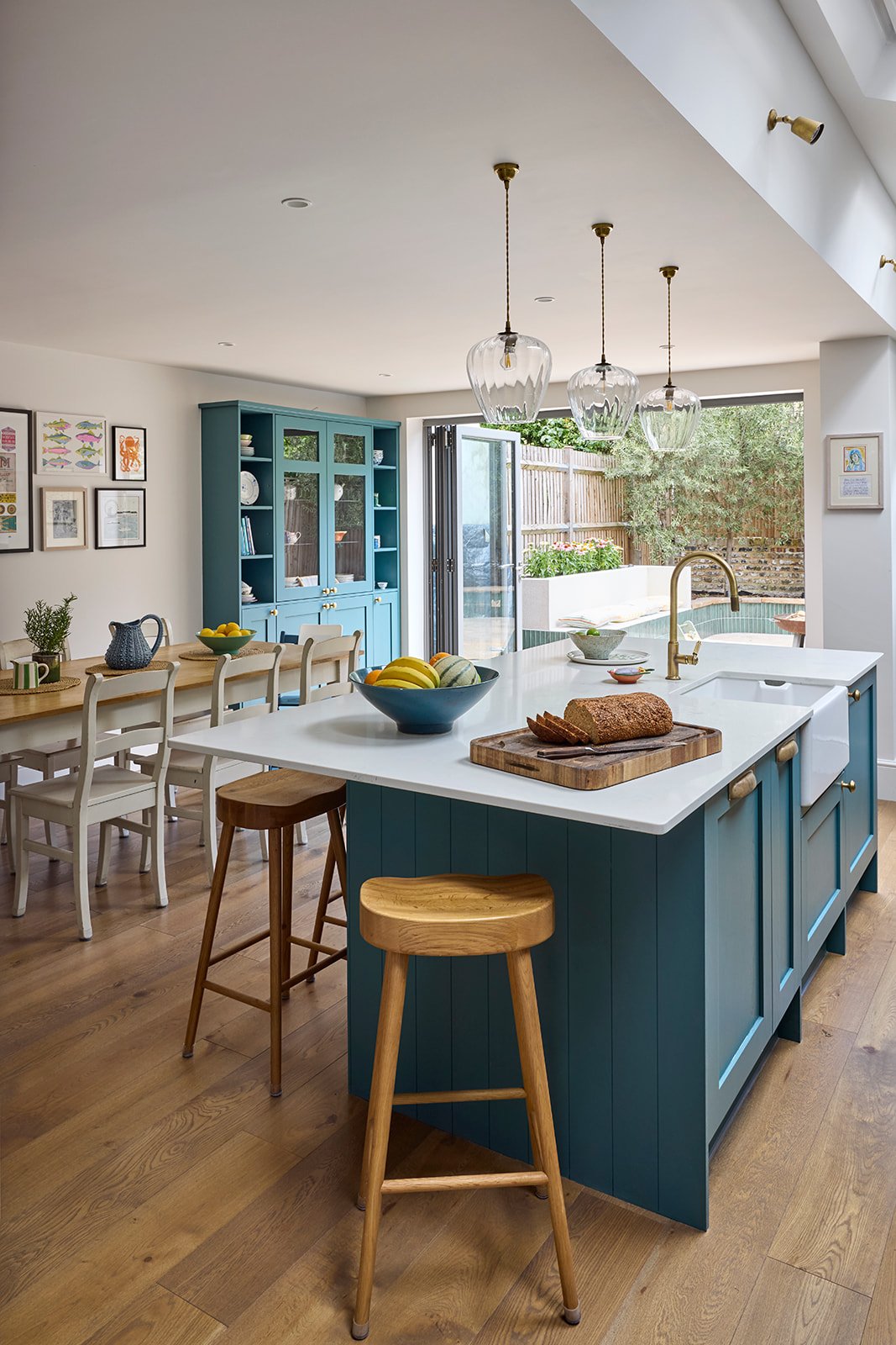 Farmhouse sink with brass finishings in a modern kitchen with teal cabinets and wooden bar stools and a dining table with a farm style chairs pendant lights teal vintage cottage display cabinet