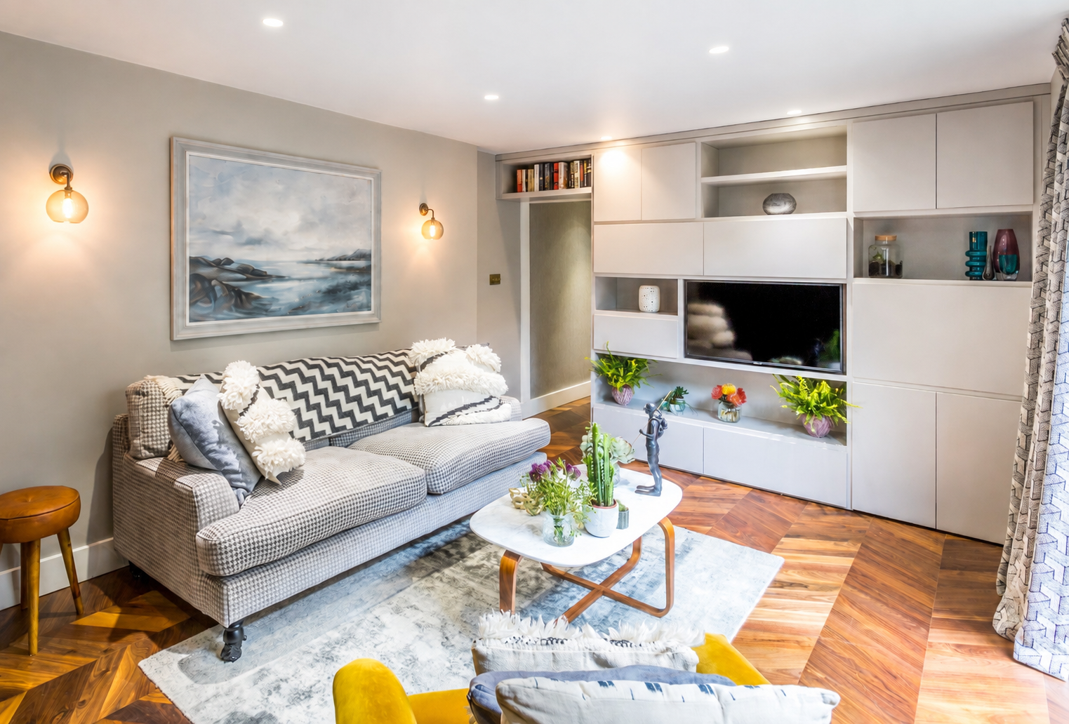 Small apartment living room with built-in storage wall, herringbone parquet floor and grey checked sofa maximising space
