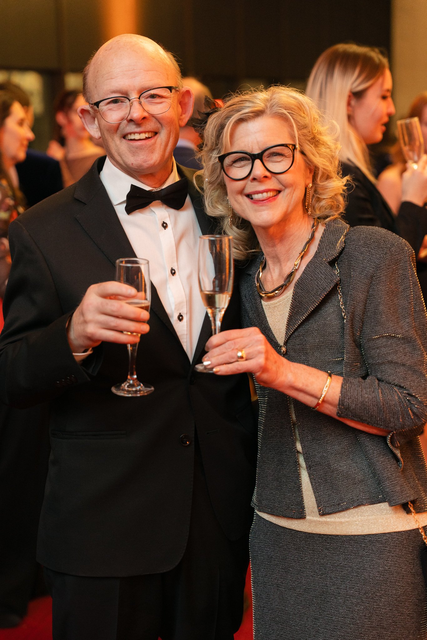 A man and woman in formal attire holding champagne glasses, smiling at an event. The man is wearing a tuxedo and the woman is in a gray suit.