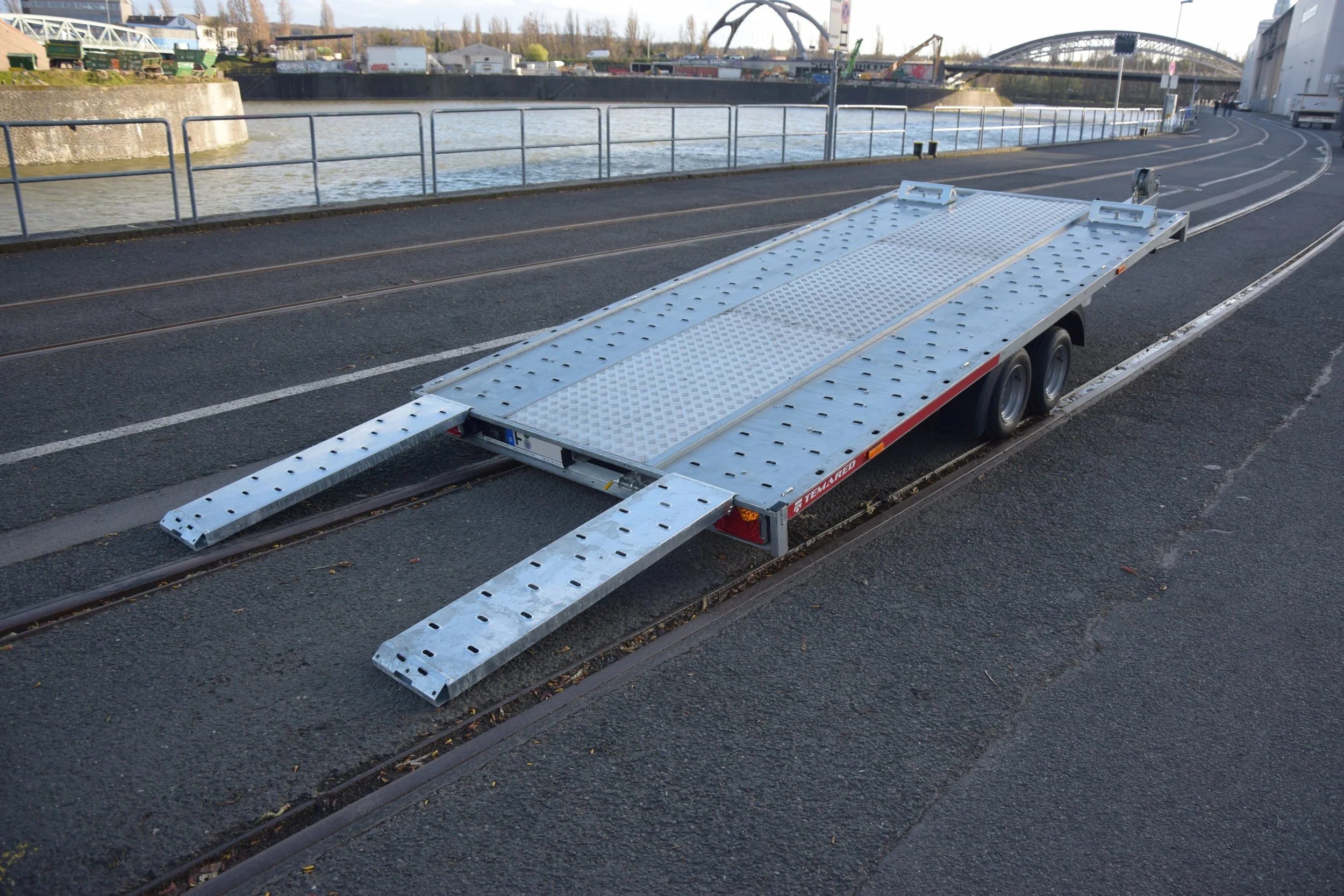 Ein leeres, silbernes Autoanhänger auf der Straße, bereit zum Transport eines Fahrzeugs, neben den Straßenbahnschienen.