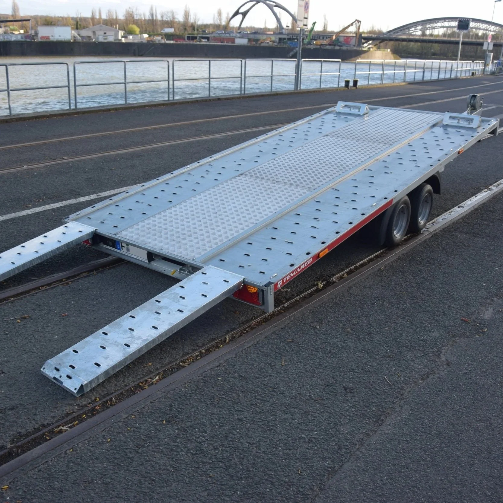Metallischer Autoanhänger mit absenkbarer Rampe, auf einer Straße mit Eisenbahnschienen, nahe einem Fluss und einer Brücke.