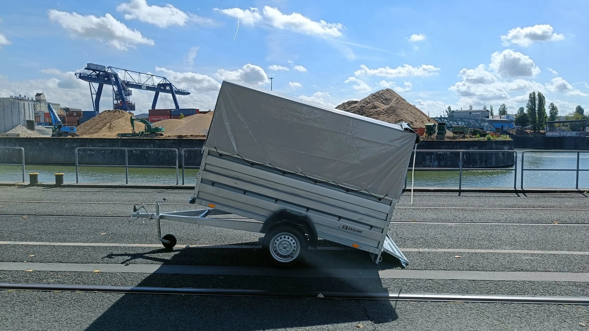 Ein kleiner, silverner Anhänger mit einem weißen Abdeckdach, der auf einem asphaltierter Weg neben Schienen steht.