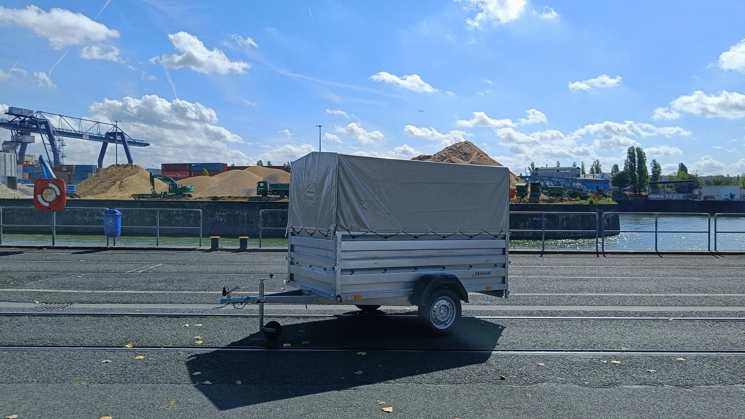 Leeres kleiner Anhänger auf einer Straße vor einem Wasserweg mit Baugeräten und Sandhaufen im Hintergrund bei sonnigem Himmel.