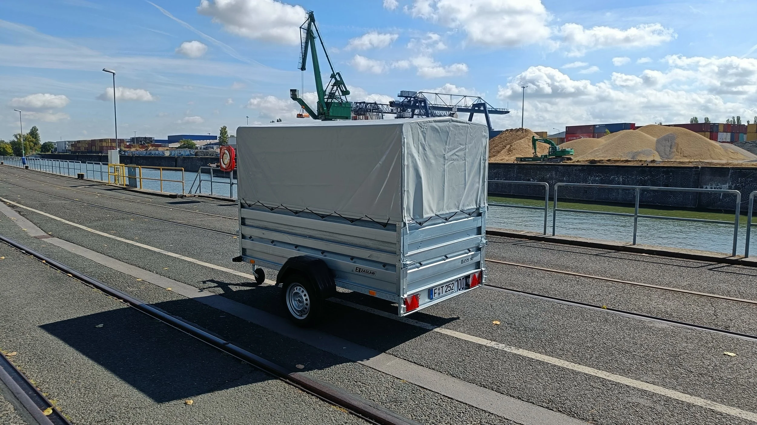 Ein kleiner, leerer Anhänger mit weißem Planenabdeckungen steht neben einer Bahnlinie an einem Wasserweg. Im Hintergrund sind Container, ein Kran und Sandhaufen sichtbar, bei sonnigem Himmel mit einigen Wolken.