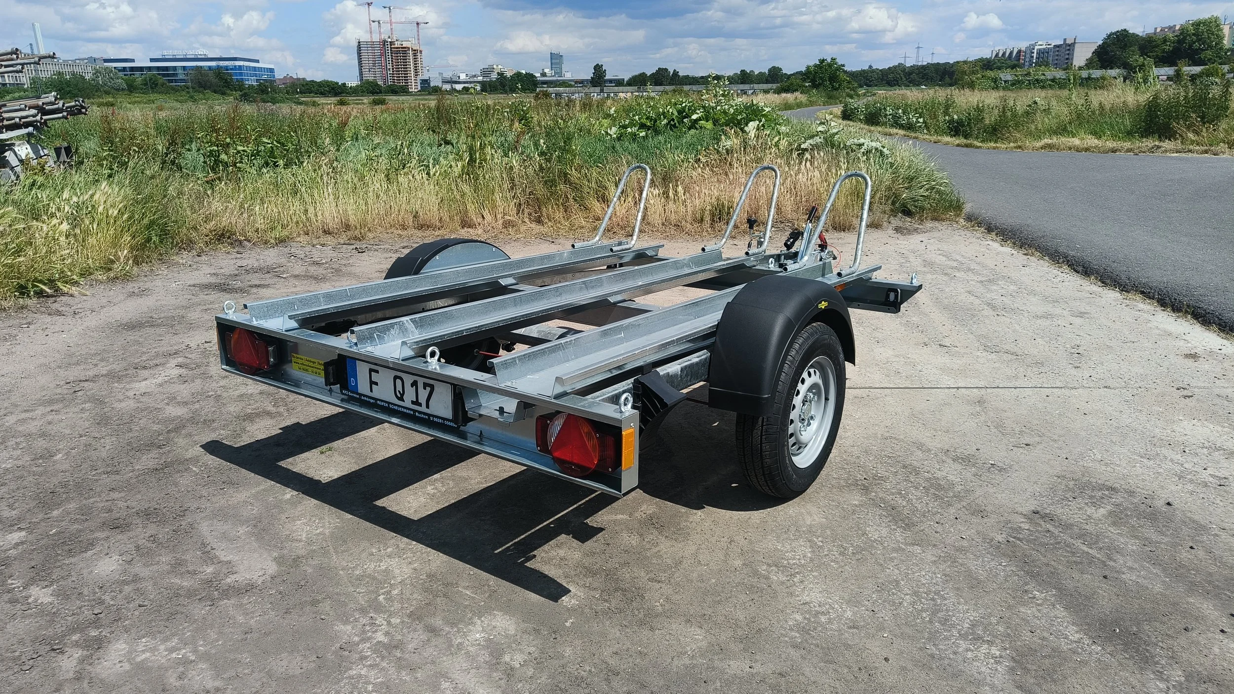 Leerer Bootstrailer auf einer unbefestigten Fläche mit Stadtgebäuden im Hintergrund.