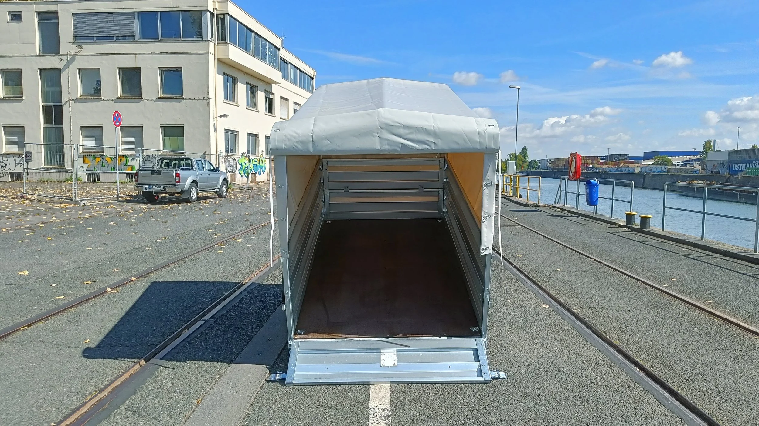 Ein offener bereitstehender Anhänger für den Transport, mit weißen Schutzdach, auf einer Uferpromenade mit Wasser, Schiffsschienen, Geländern, Graffiti-Gebäude im Hintergrund und einem blauen Himmel.