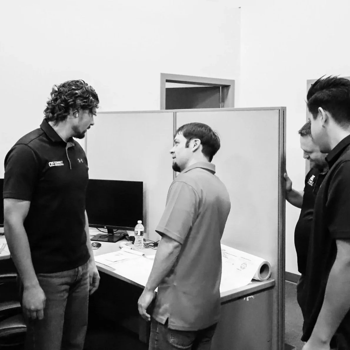 Four men standing in an office or conference room, engaged in conversation, with a computer monitor, water bottle, and some papers on a desk. Coworkers at Connect Engineering, a civil engineering and land surveying business.