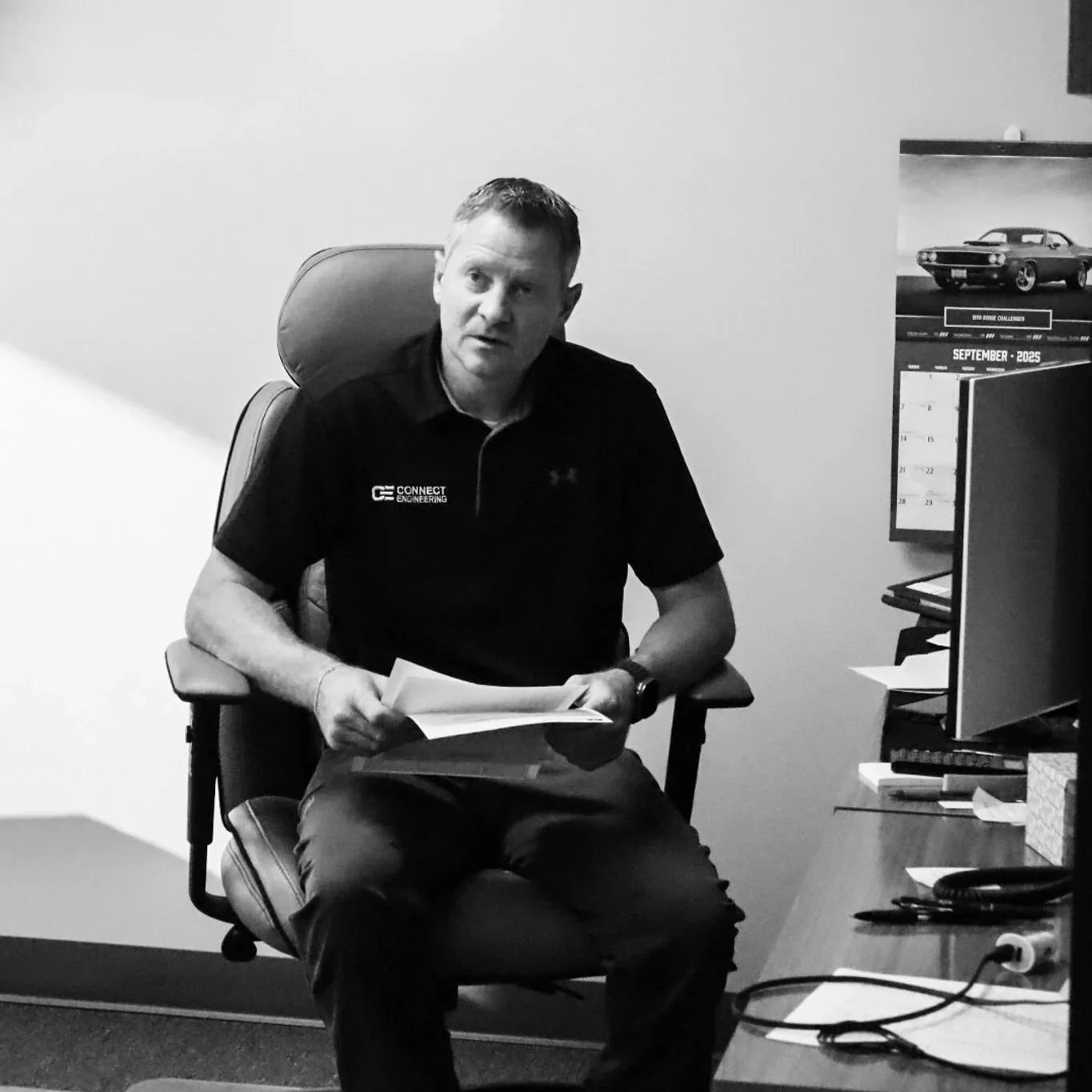 Black-and-white photo of a man sitting in an office chair, holding papers, with a desk and calendar visible in the background. Blake Jolley with Connect Engineering and Surveying