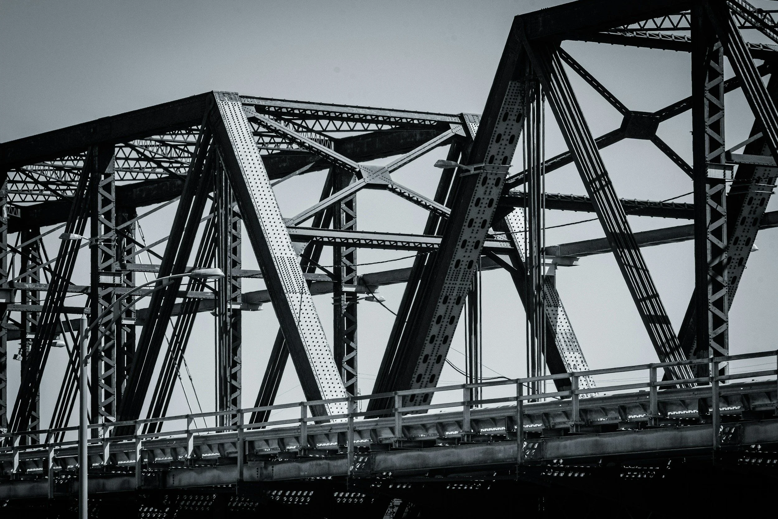 Black and white photo of a steel bridge structure with intersecting beams and a railing at the bottom.