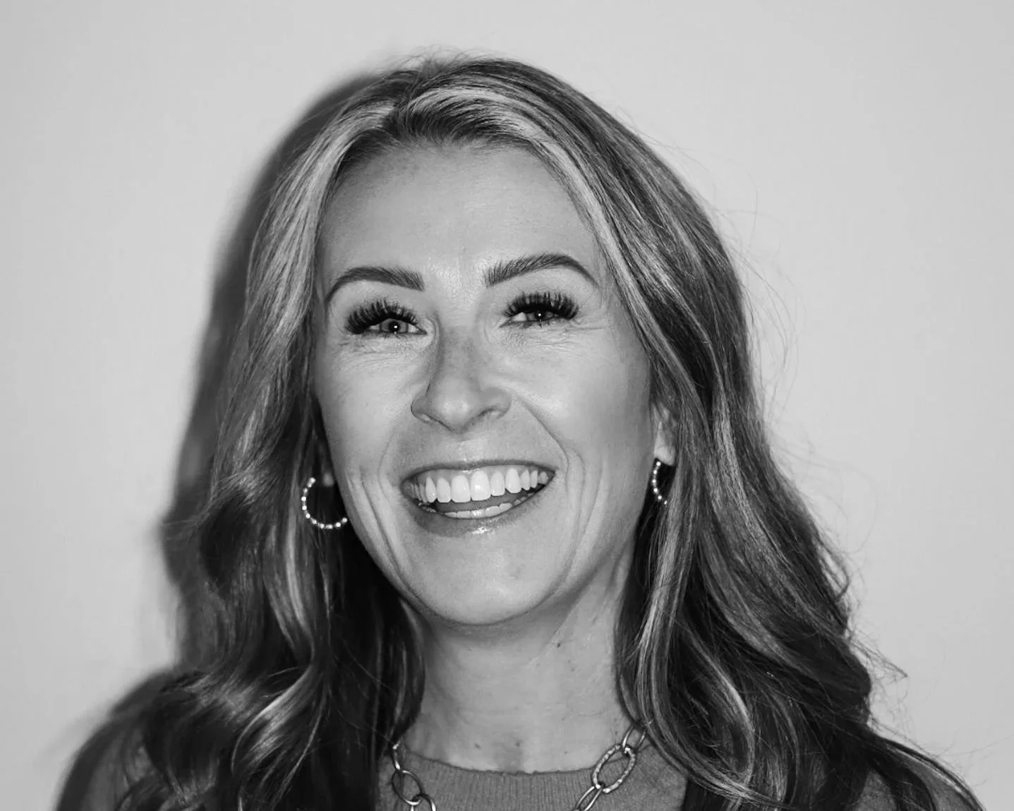 Black and white portrait of a smiling woman with long wavy hair, wearing hoop earrings and a chain necklace with a pendant. A Connect Engineering Employee.