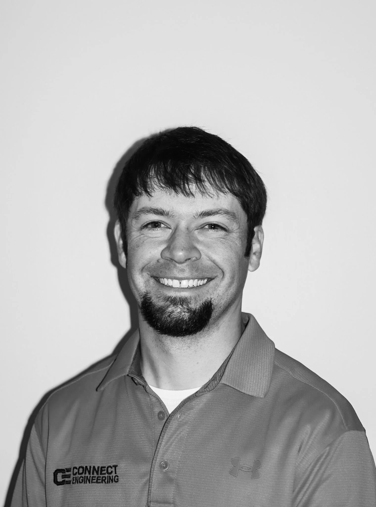 Black and white headshot of a man smiling, wearing a polo shirt with the logo 'Connect Engineering' and Under Armour branding. A Connect Engineering Employee.