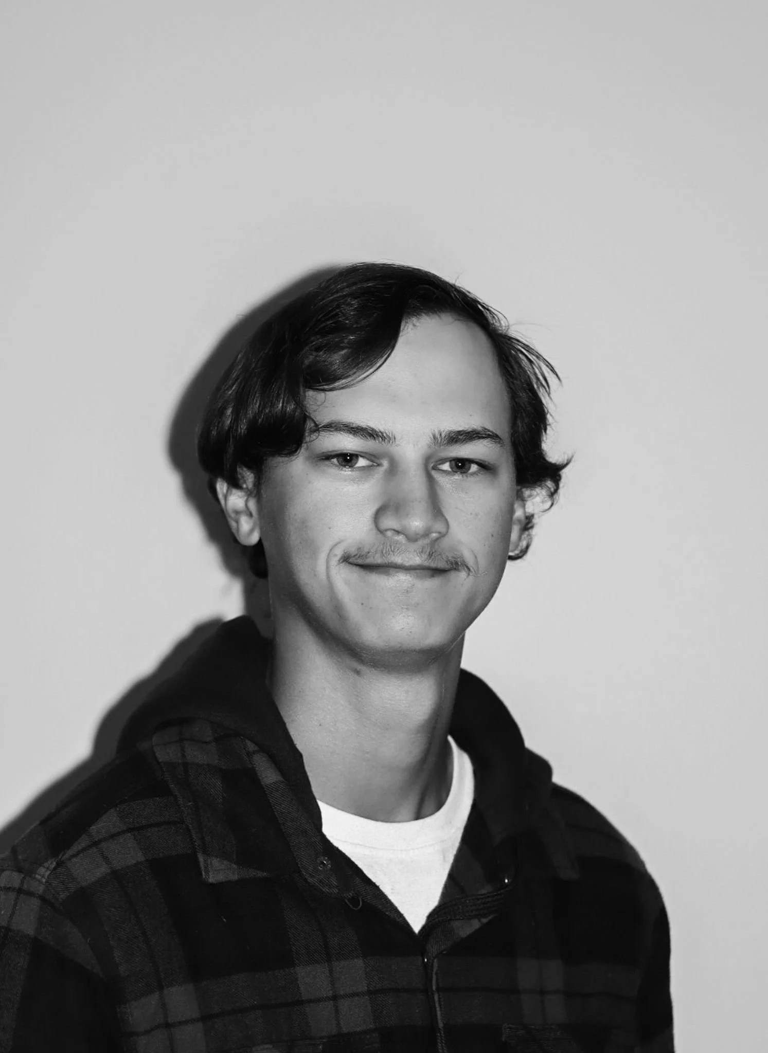 Black and white portrait of a young man with dark hair, a mustache, wearing a checkered jacket with a hoodie, standing against a plain background. A Connect Engineering Employee.