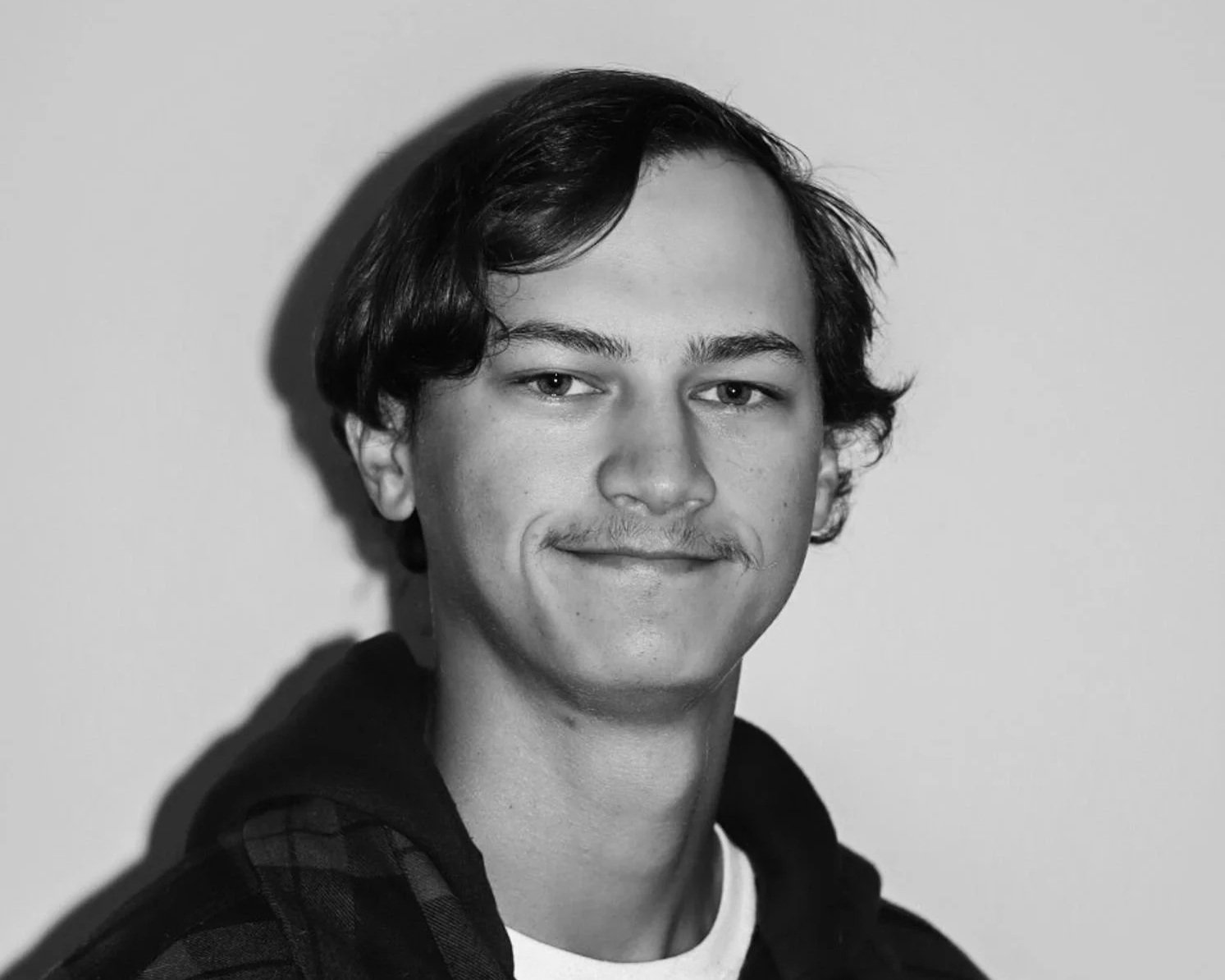 Black and white photo of a young man with medium-length dark hair, light facial hair, and a slight smile, wearing a checked shirt with a hooded sweatshirt underneath, standing against a plain background. A connect Engineering employee. 