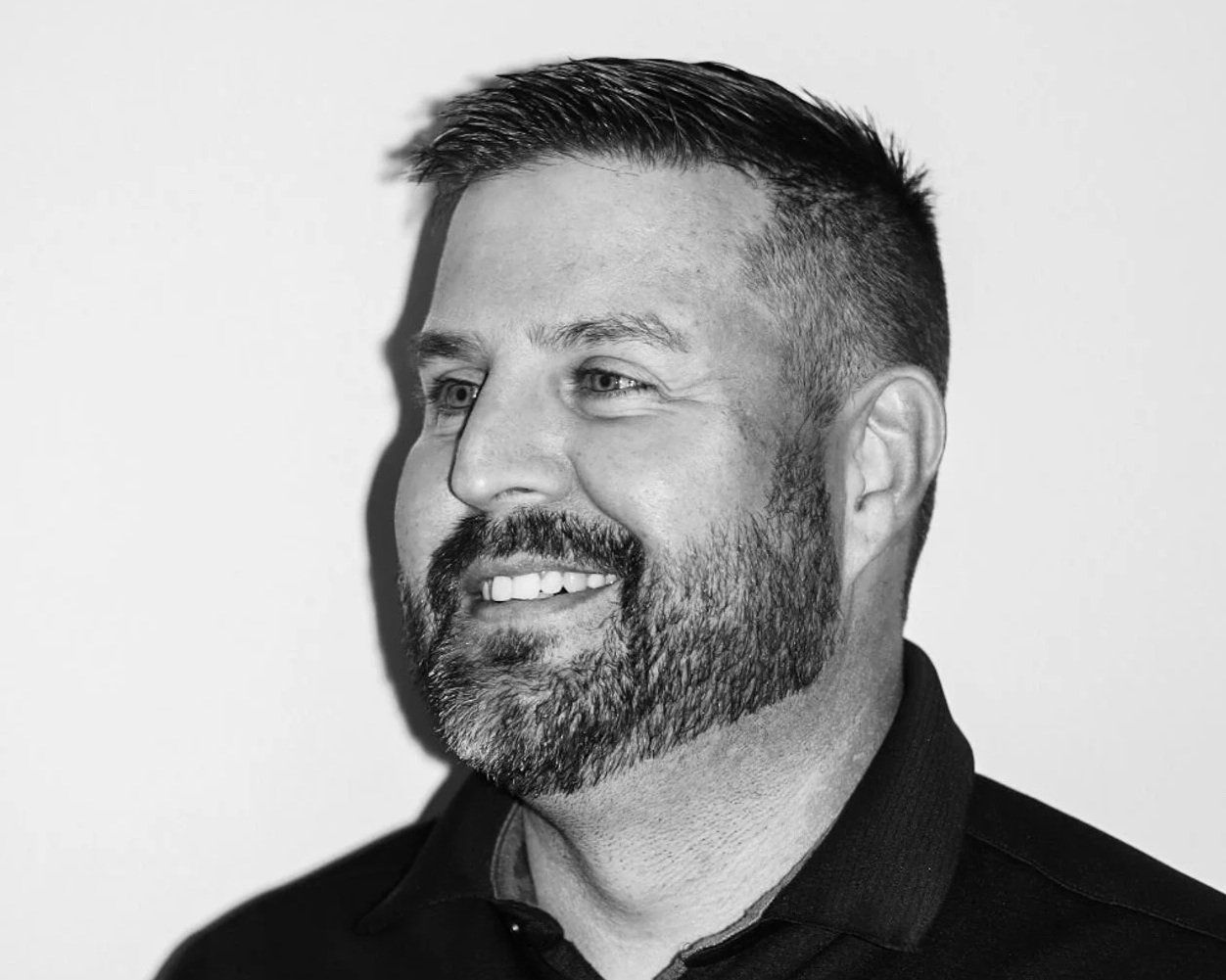 Black and white photo of a smiling man with a beard and short hair, wearing a dark collared shirt. A Connect Engineering Employee.