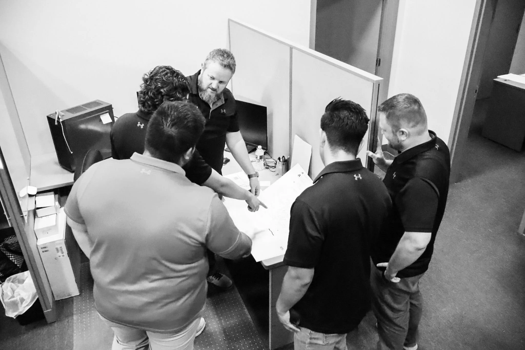 Six men gathered around a desk in an office, looking at documents and discussing projects for civil engineering, land entitlements, land surveying, construction quality assurance and quality control, master planning.