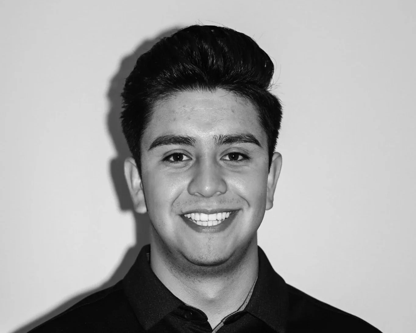 Black and white portrait of a young man with dark hair, smiling, wearing a dark shirt, standing against a plain light background. A Connect Engineering Employee.
