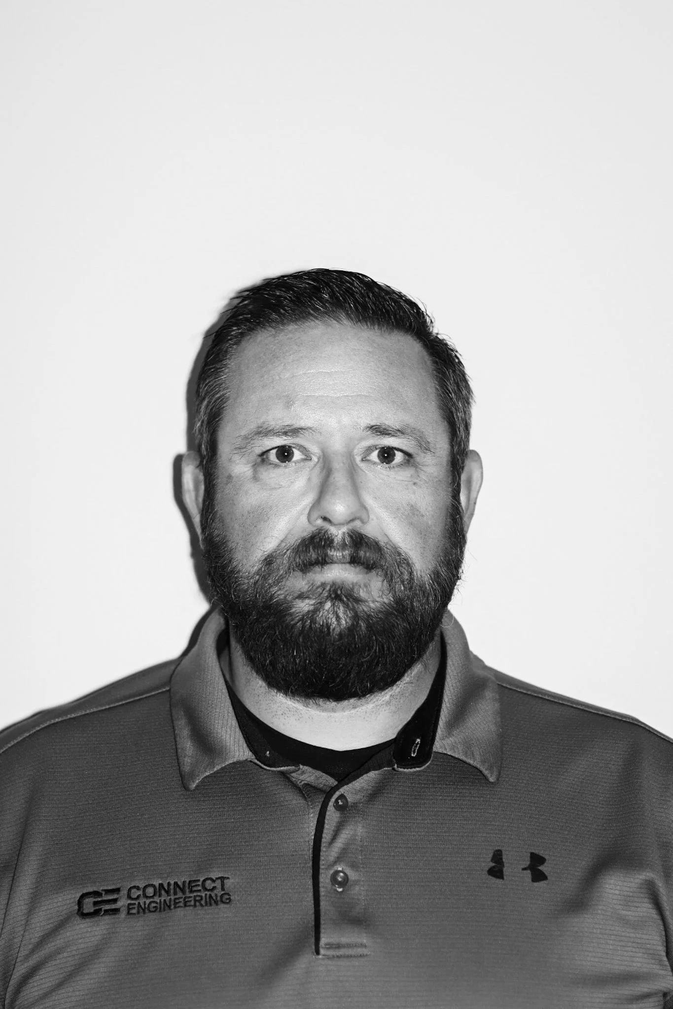 Black and white portrait of a man with a beard, wearing a collared shirt with the logo 'Connect Engineering' and Under Armour logo. A Connect Engineering Employee.