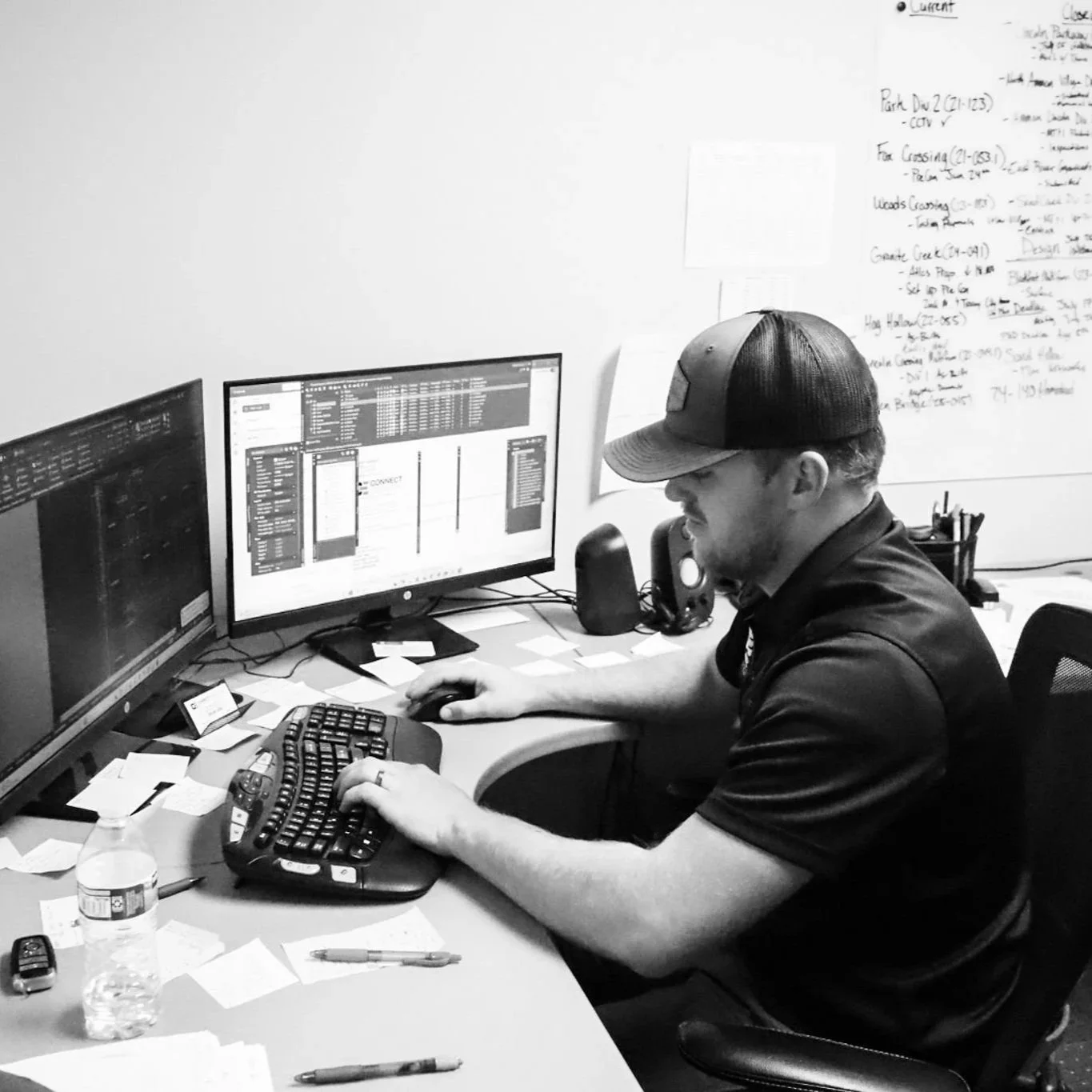 A man wearing a cap and glasses sitting at a cluttered desk with two monitors, using a computer keyboard and mouse, with handwritten notes and sticky notes on the desk.