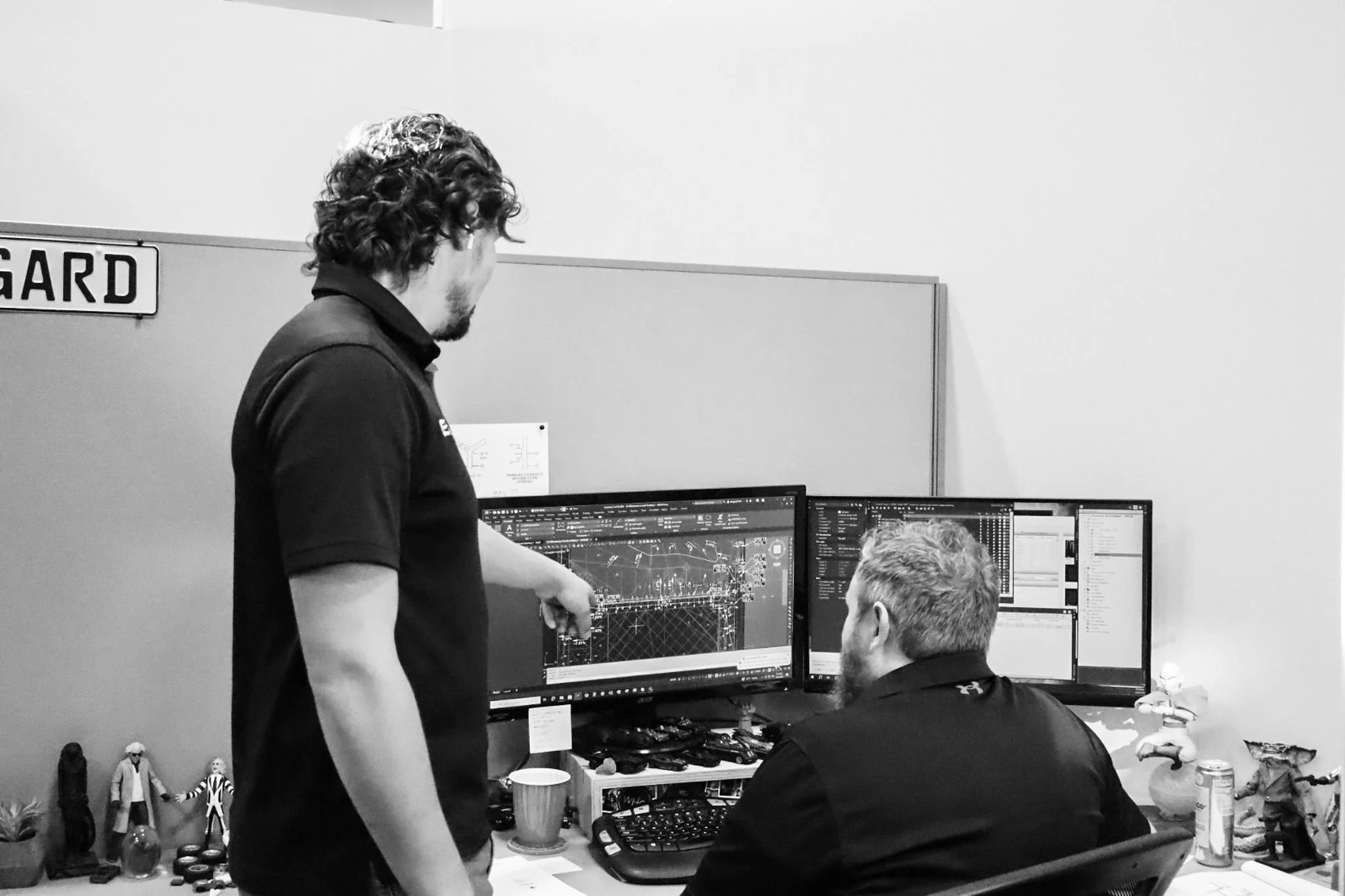 Two men are working at a desk with three computer monitors showing technical diagrams for civil engineering projects. One man is standing, pointing at one monitor, while the other is seated, looking at the screens.