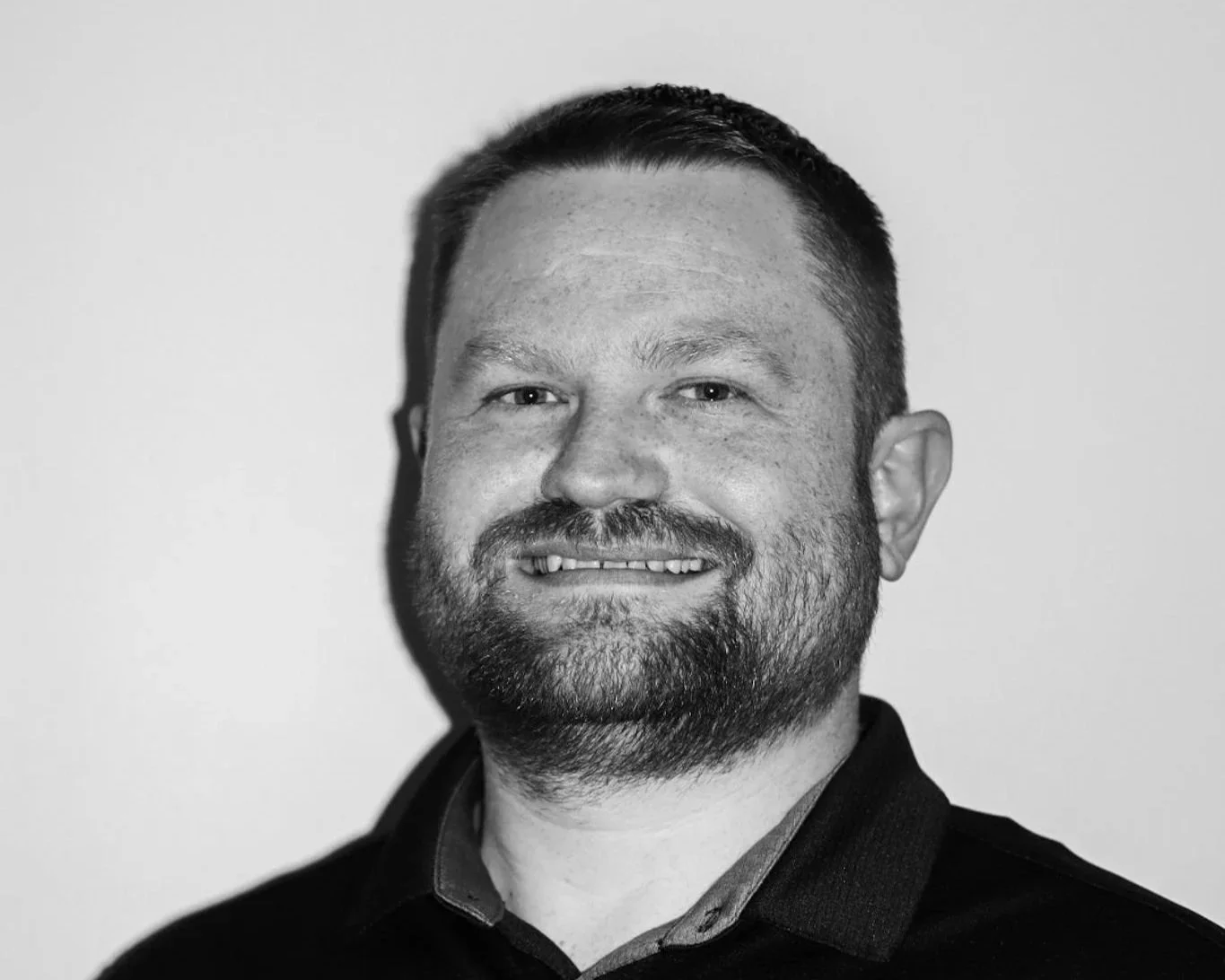 Black and white portrait of a man with short hair and a beard, smiling slightly, wearing a collared shirt and a jacket. A Connect Engineering Employee.