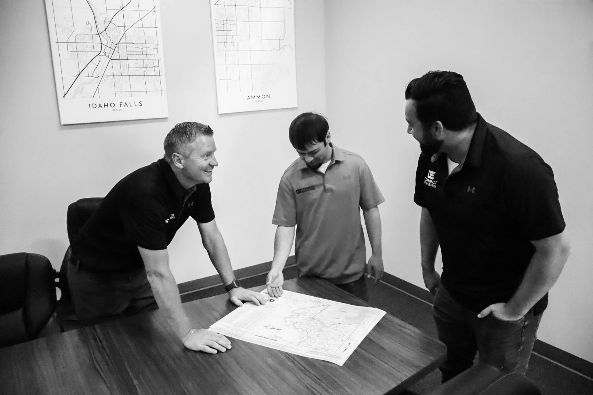 Three men and a boy in a meeting room looking at a civil engineering and land surveying plans on a table, with two maps on the wall behind them labeled 'Idaho Falls' and 'Ammon'.